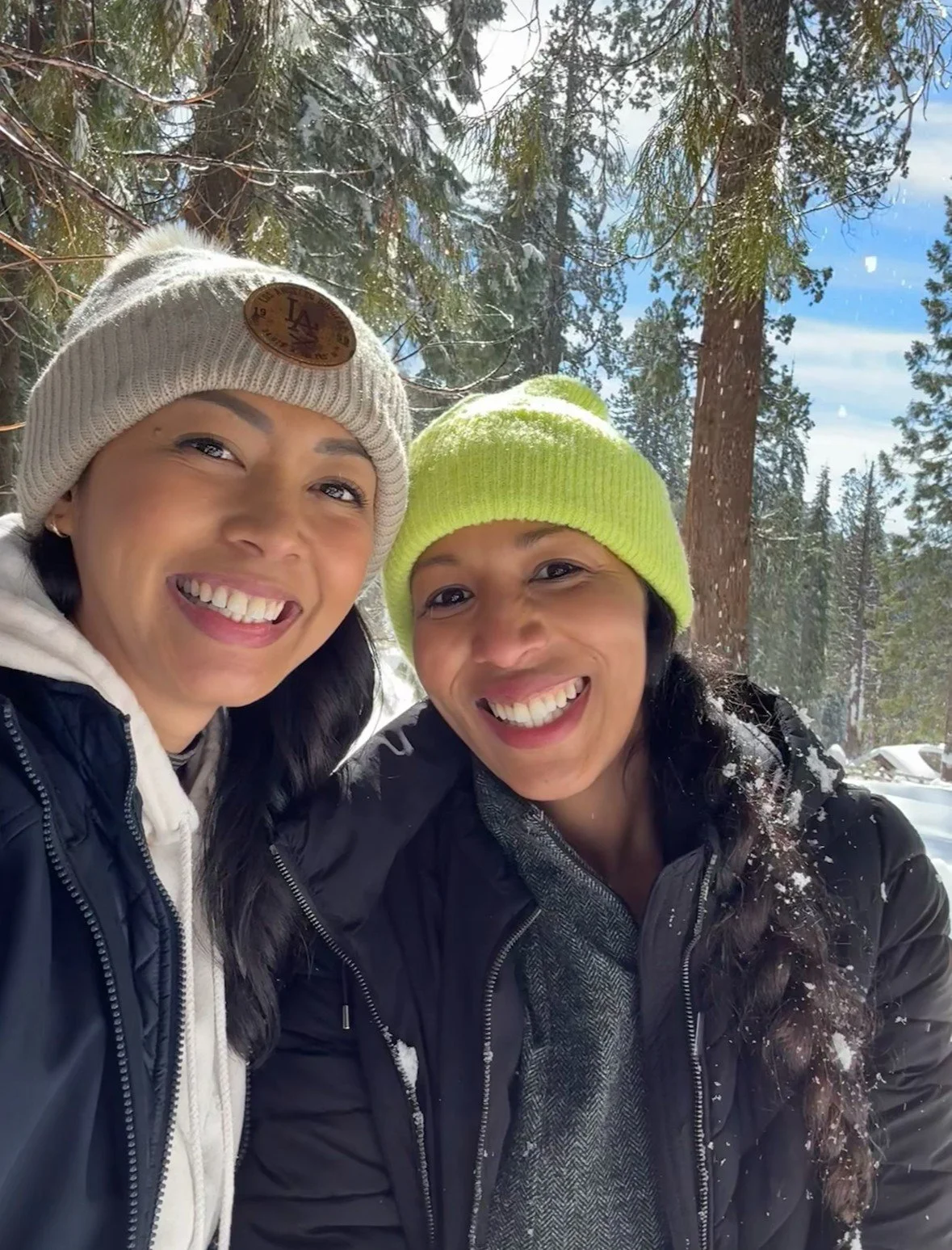 Two women smiling in a snowy forest with trees and a blue sky in the background, wearing winter hats and jackets.