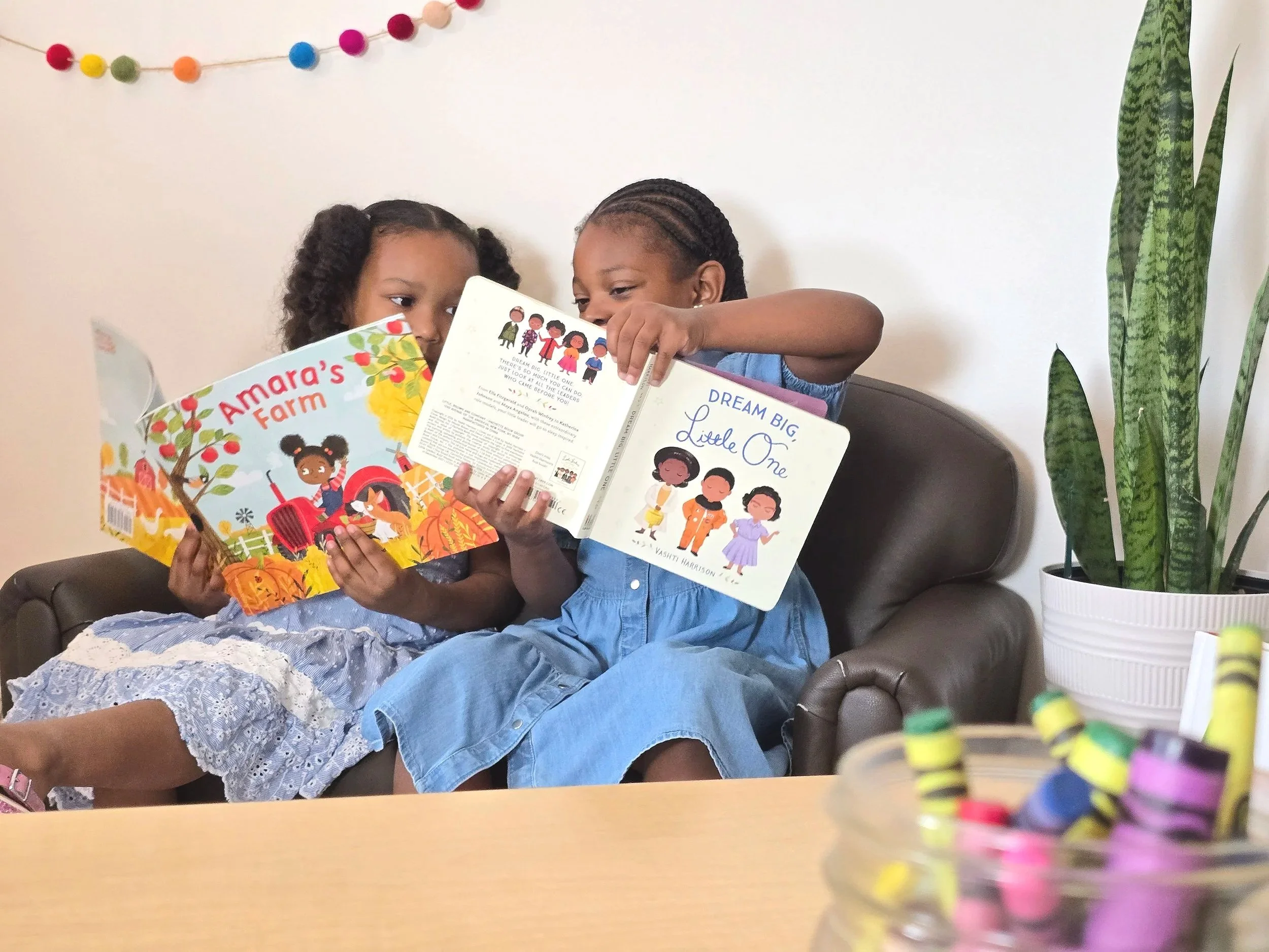 Two young girls sitting on a brown leather couch, reading books together. The girl on the left has curly hair and is wearing a white lace dress, while the girl on the right has braided hair and is dressed in a blue shirt. There is a tall green plant on the right side of the image and a garland of colorful pom-poms hanging on the white wall in the background.