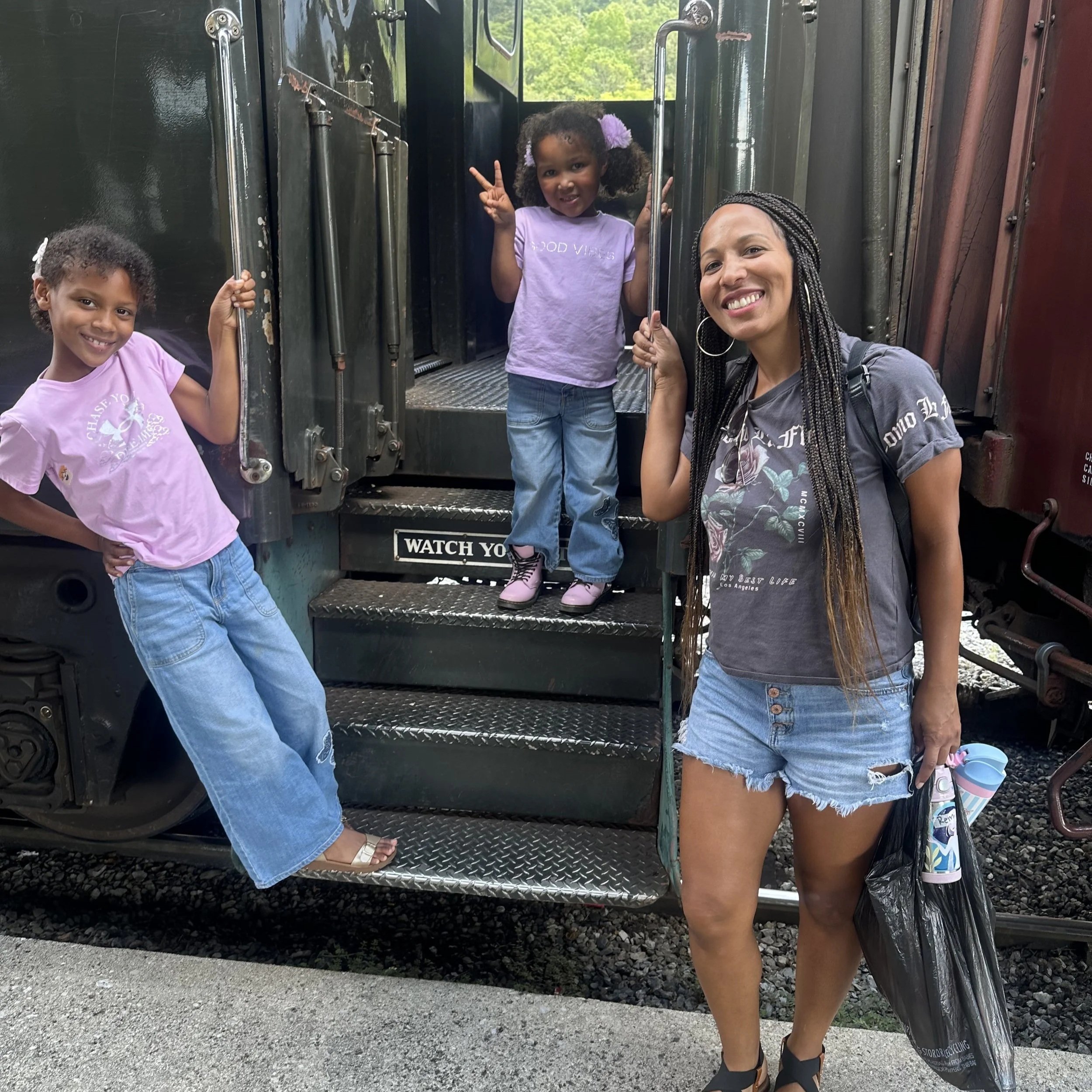Three girls and one woman standing in front of a train, holding onto the train's handrails, with green trees in the background. One girl is smiling with her hand on her hip, another girl is making a peace sign, the woman is smiling, and the third girl is standing at the train's door with one hand up.