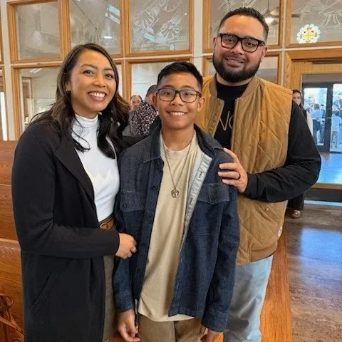 A family of three posing together indoors, smiling at the camera. The woman is on the left, the young boy in the middle, and the man is on the right, with wooden decor and windows in the background.