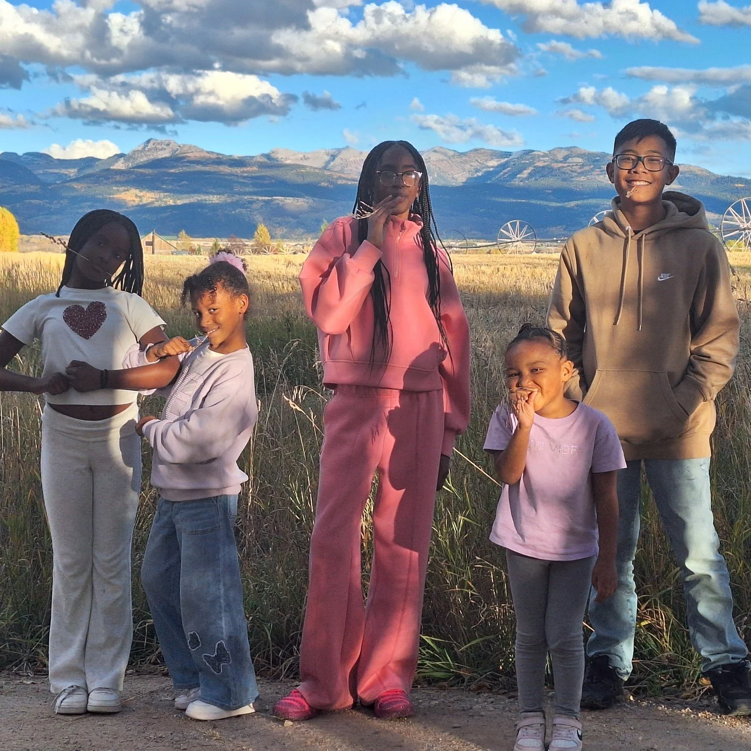 A group of six children standing outdoors in a field with mountains and blue sky with clouds in the background.