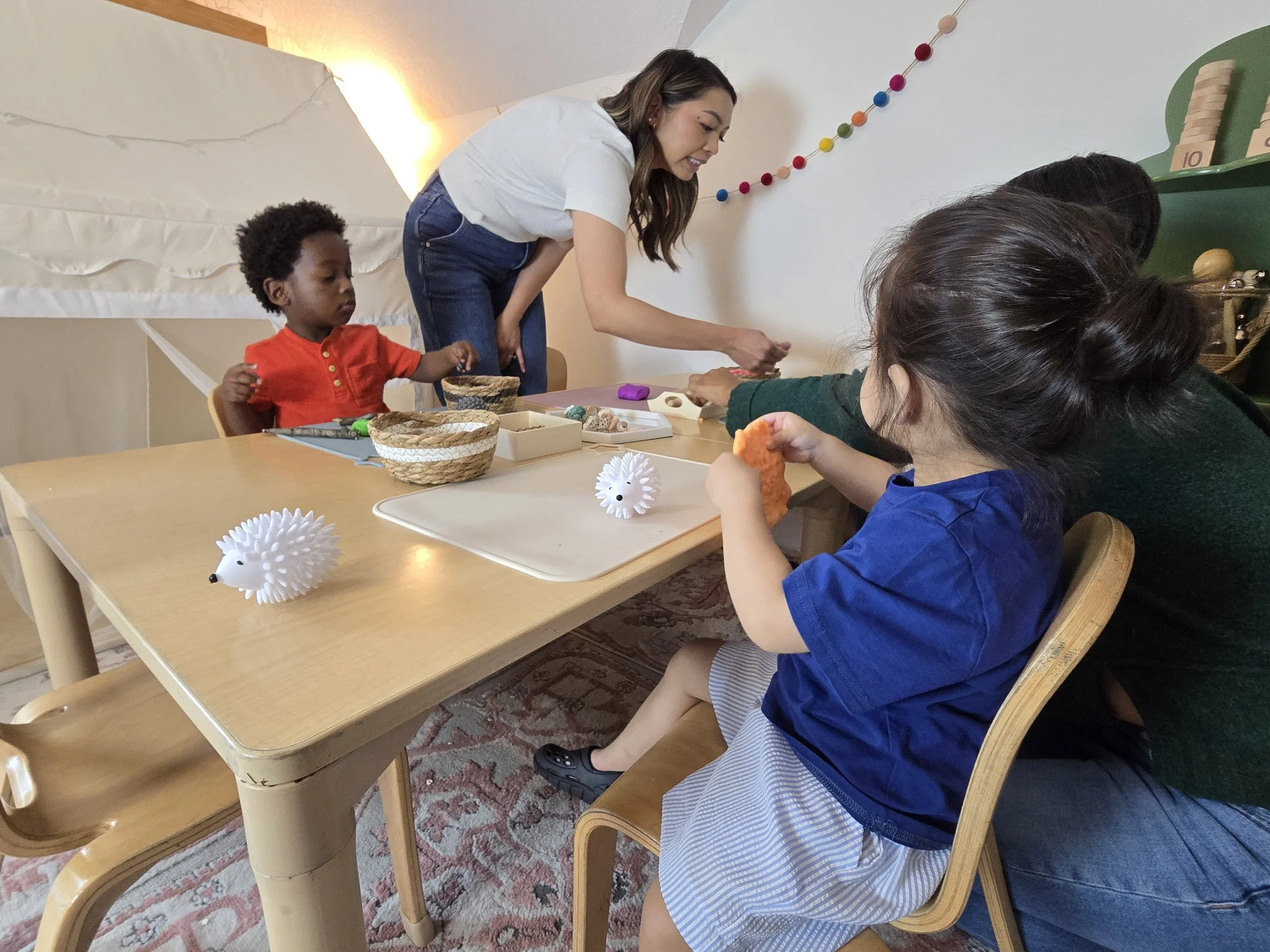 A woman showing children craft supplies at a table decorated with hedgehog-shaped toys and surrounded by colorful decorations on the wall.