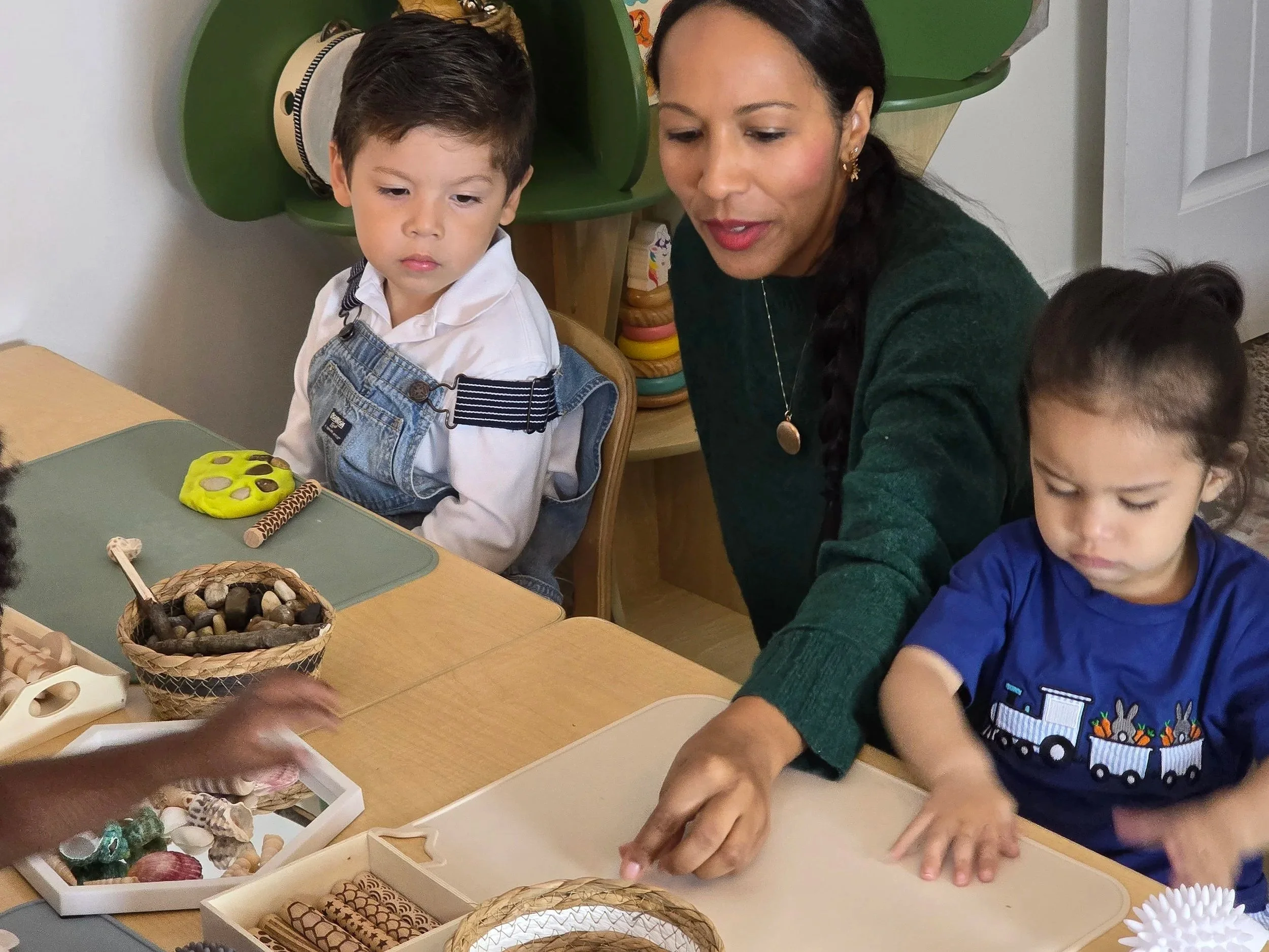 A woman with dark hair, wearing a dark green sweater, and two young boys are sitting at a table engaging in a crafting activity. The boy on the left has short dark hair and is wearing white shirt with denim overalls. There are various craft materials and baskets on the table.
