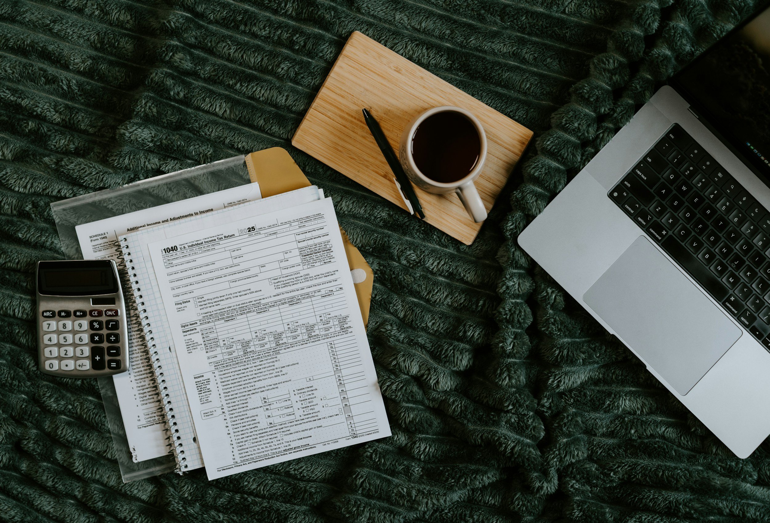 A workspace with a laptop, a coffee mug, a pen, a calculator, and filled-out tax forms on a dark green textured blanket.