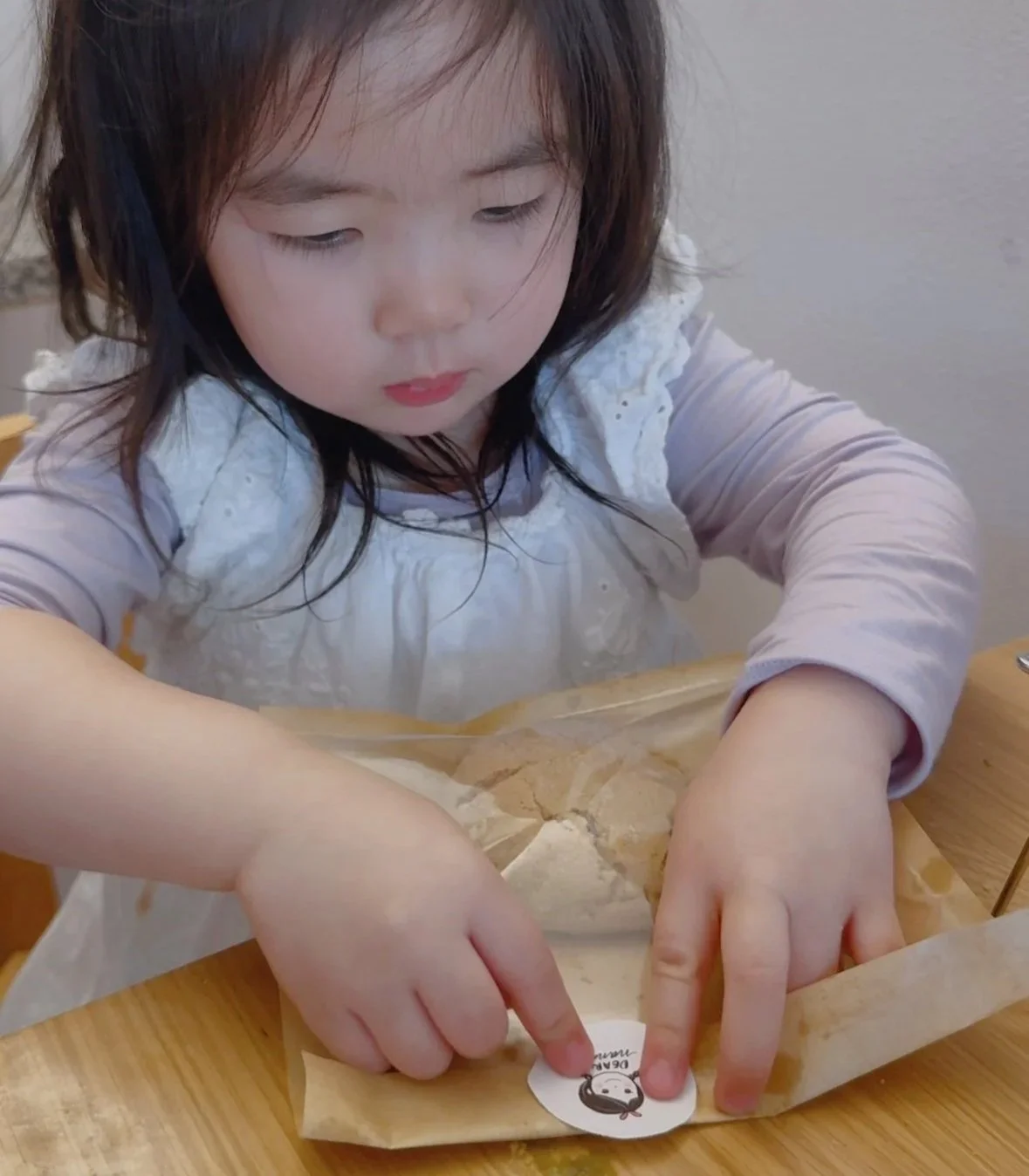 A young girl with dark hair is unwrapping a pastry or bread from brown parchment paper at a wooden table.