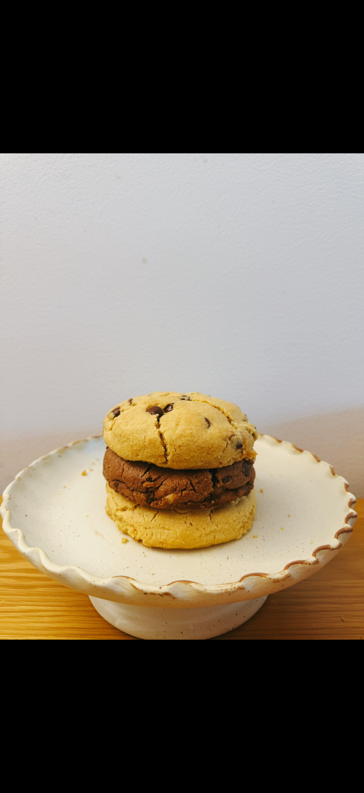 Close-up of a plate with three cookies stacked on top of each other, placed on a wooden surface against a plain wall background.