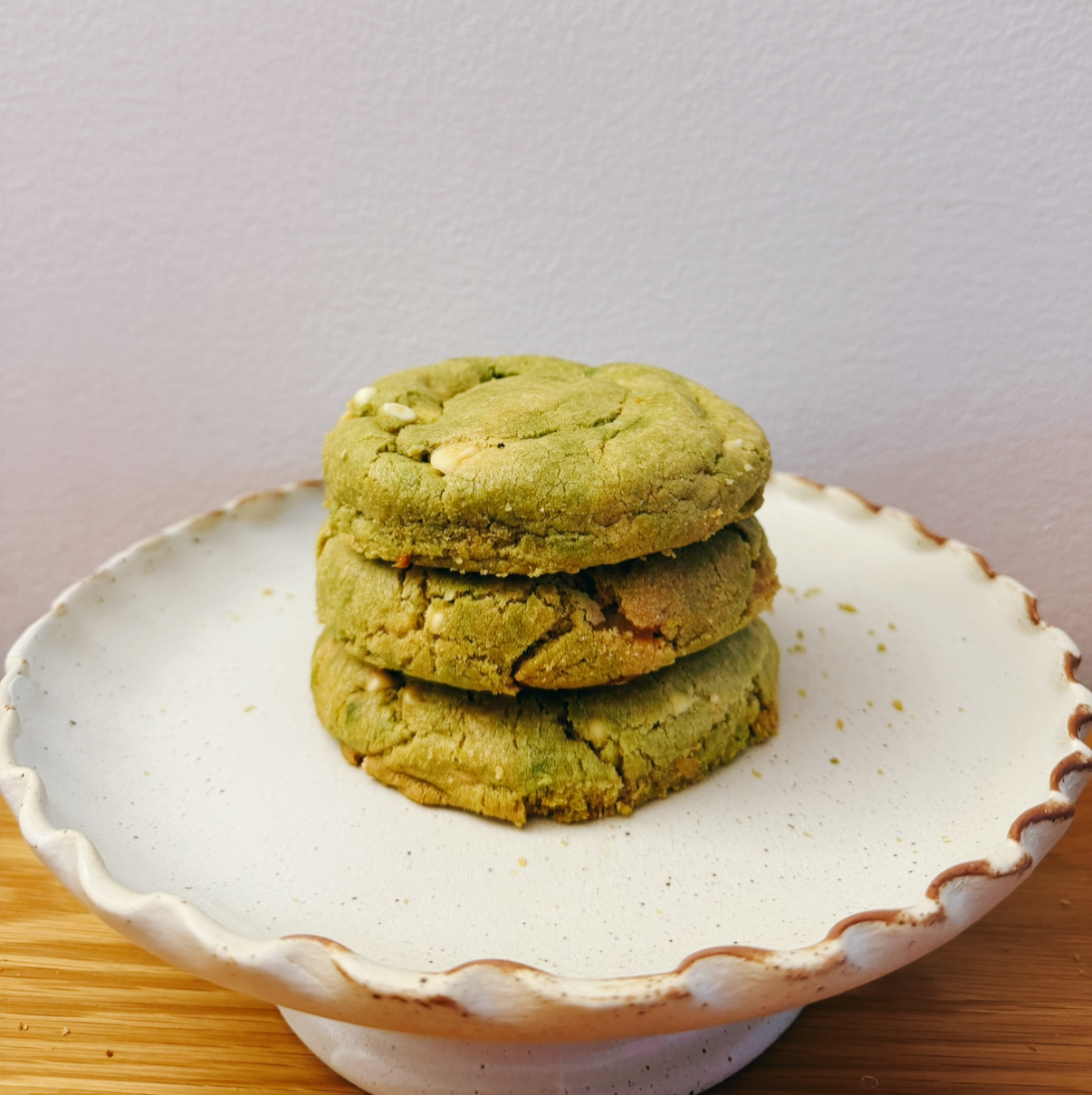 Three green cookies stacked on a white ceramic plate with a scalloped edge, placed on a wooden surface.
