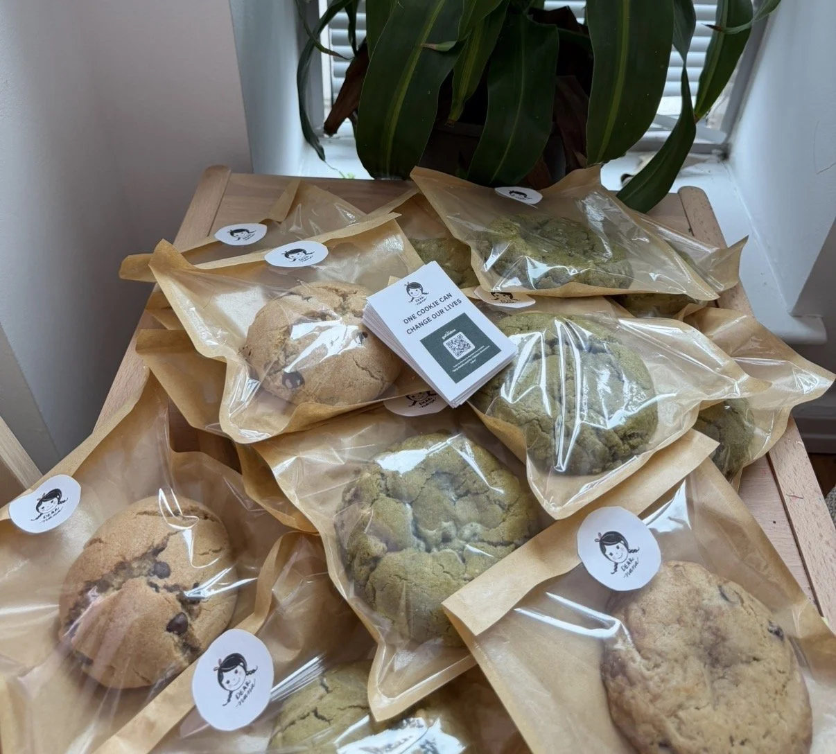 Assortment of packaged cookies on a wooden table near a window with green plant in the background.