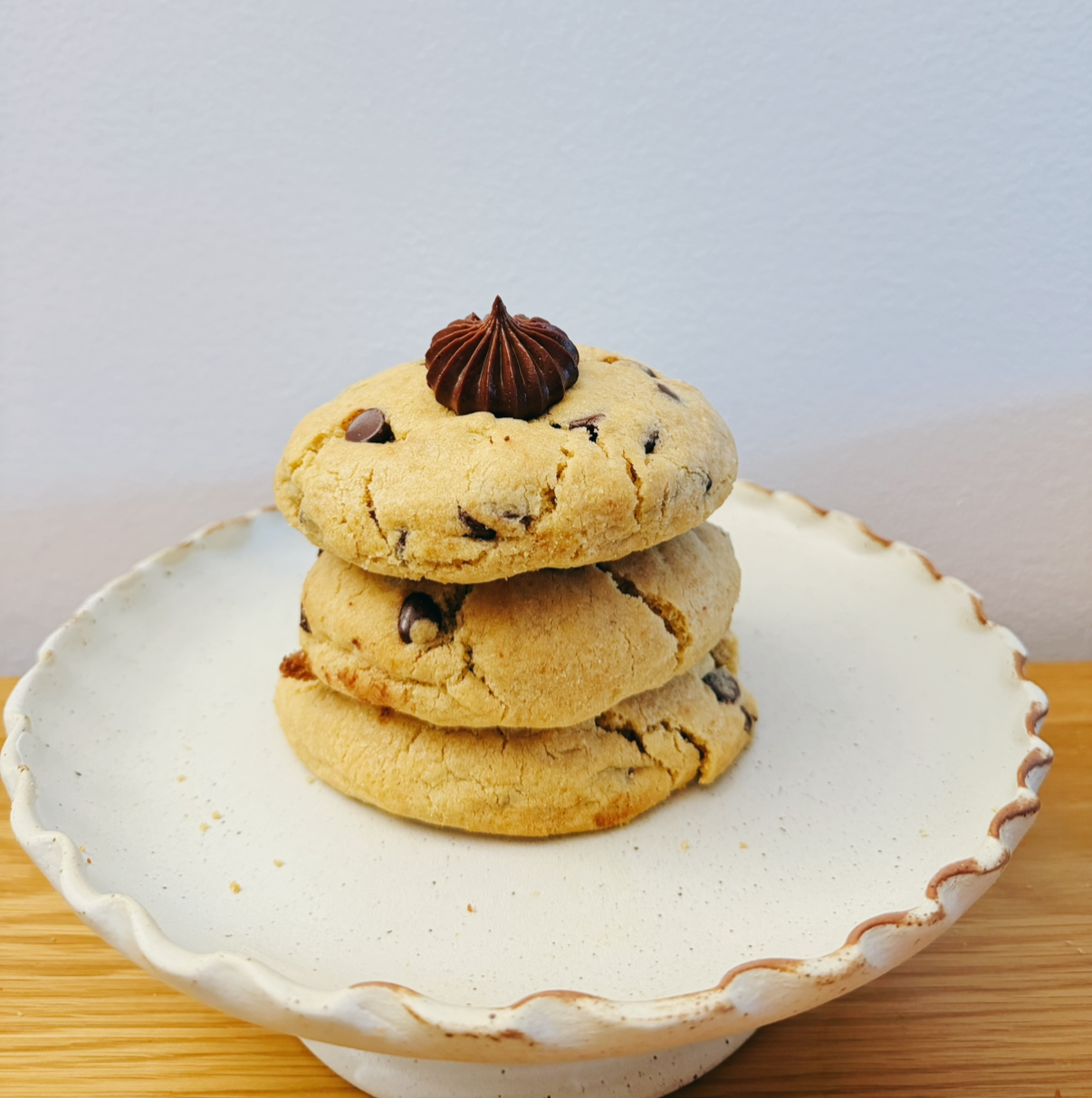 Three chocolate chip cookies stacked on a white plate with a decorative rim, topped with a chocolate swirl, on a wooden surface against a plain wall.