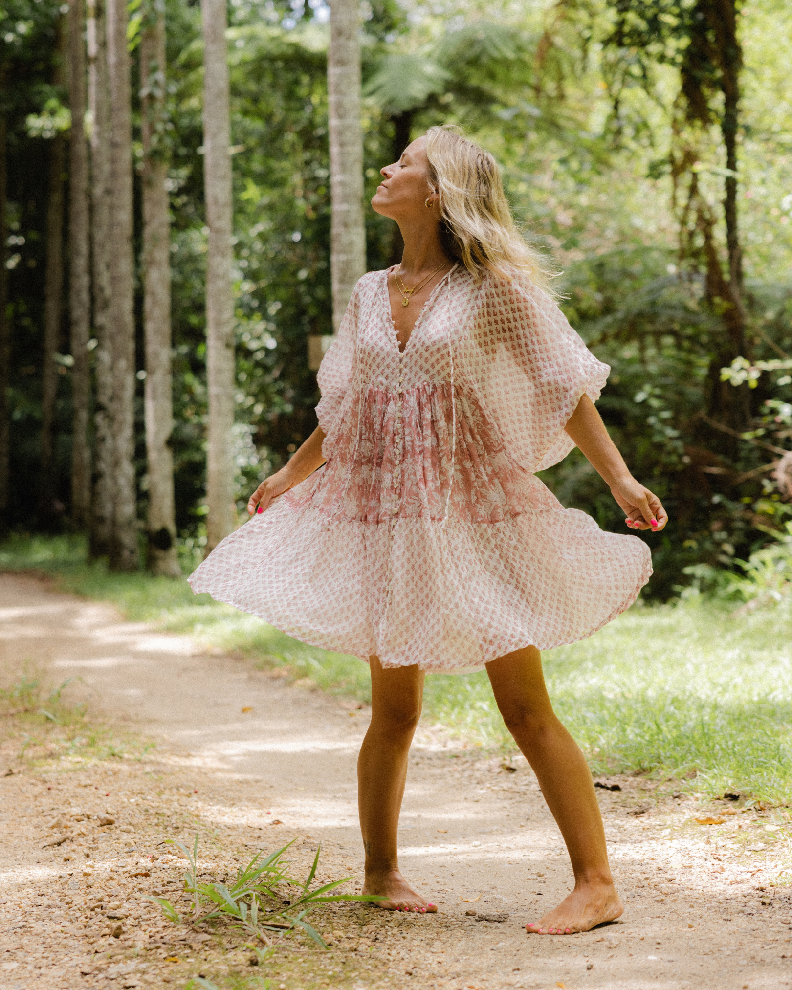 A woman in a flowing pink dress standing barefoot on a dirt path in a green forest, with her eyes closed and hands stretched outward.