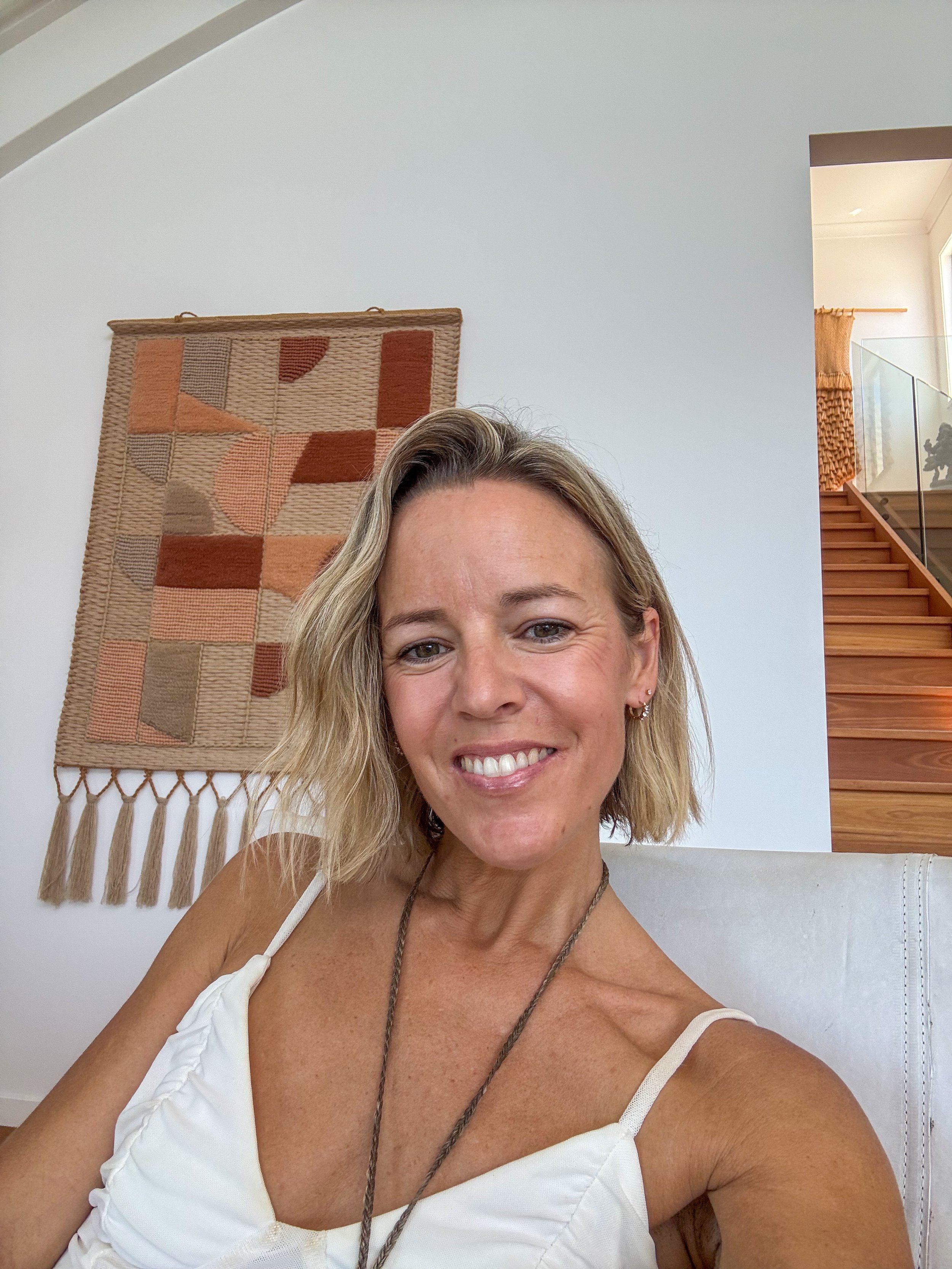 Smiling woman with short blonde hair sitting in a modern home interior with a colorful woven wall hanging and wooden staircase in the background.
