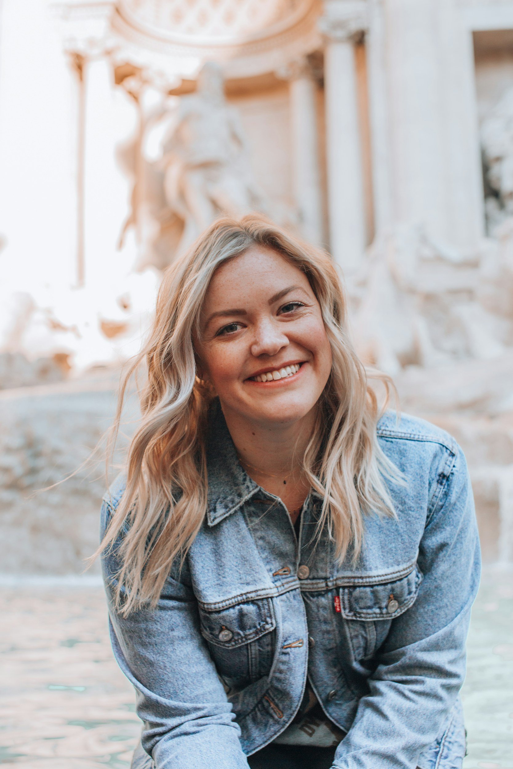 A young woman with blonde wavy hair smiling outdoors in front of a classical statue and stone architecture.