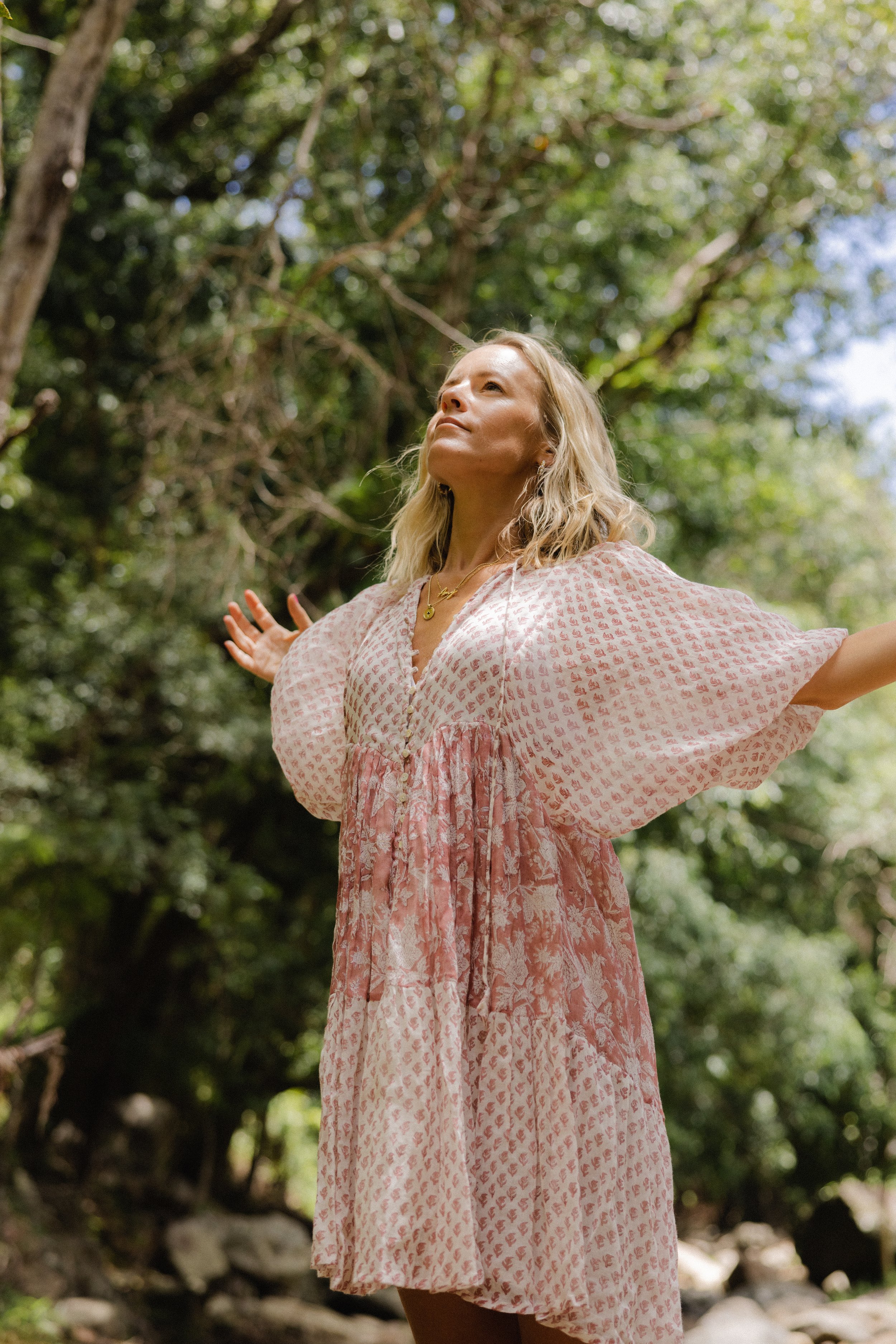 Woman with blonde hair wearing a pink and white patterned dress standing outdoors with arms outstretched in a lush green forest, looking up.
