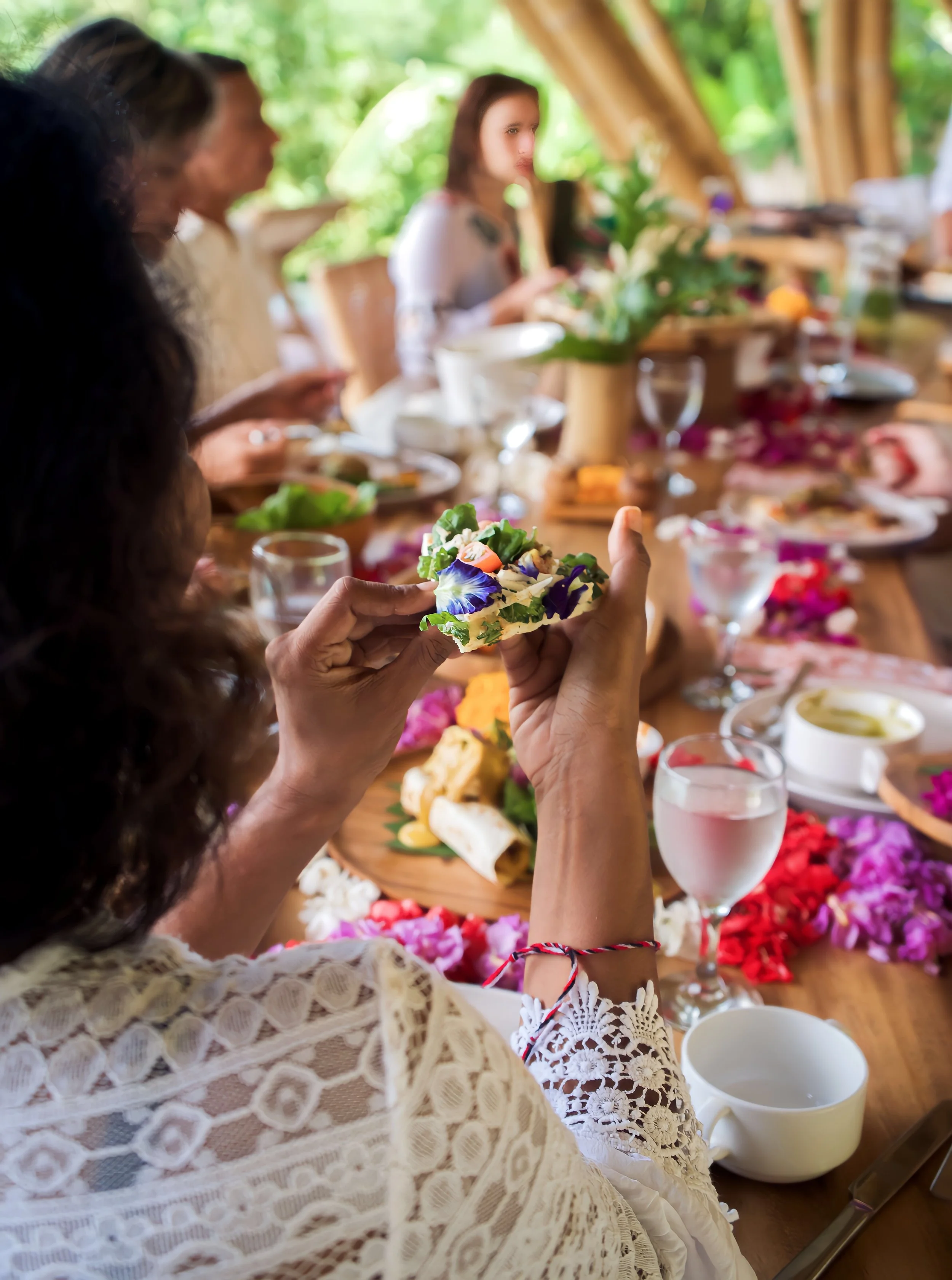 People at a dining table in a bright, outdoor setting, with a focus on a person holding a decorated piece of food or napkin, surrounded by various dishes and drinks, colorful flower petals on the table.