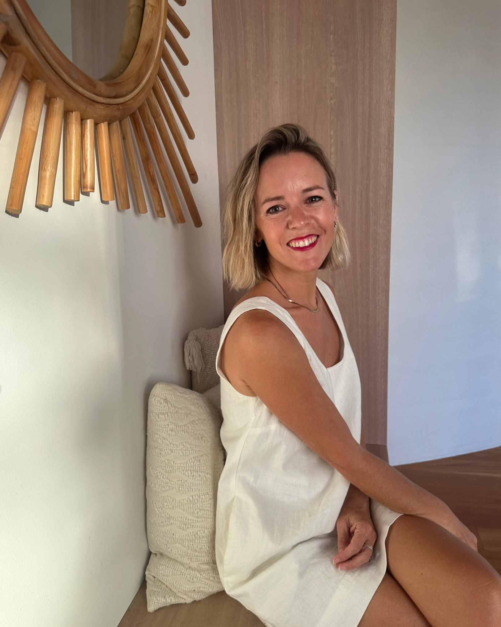 A woman sitting on a wooden floor with a cushion behind her, smiling at the camera, wearing a white sleeveless dress, with a wooden wall and mirror behind her.