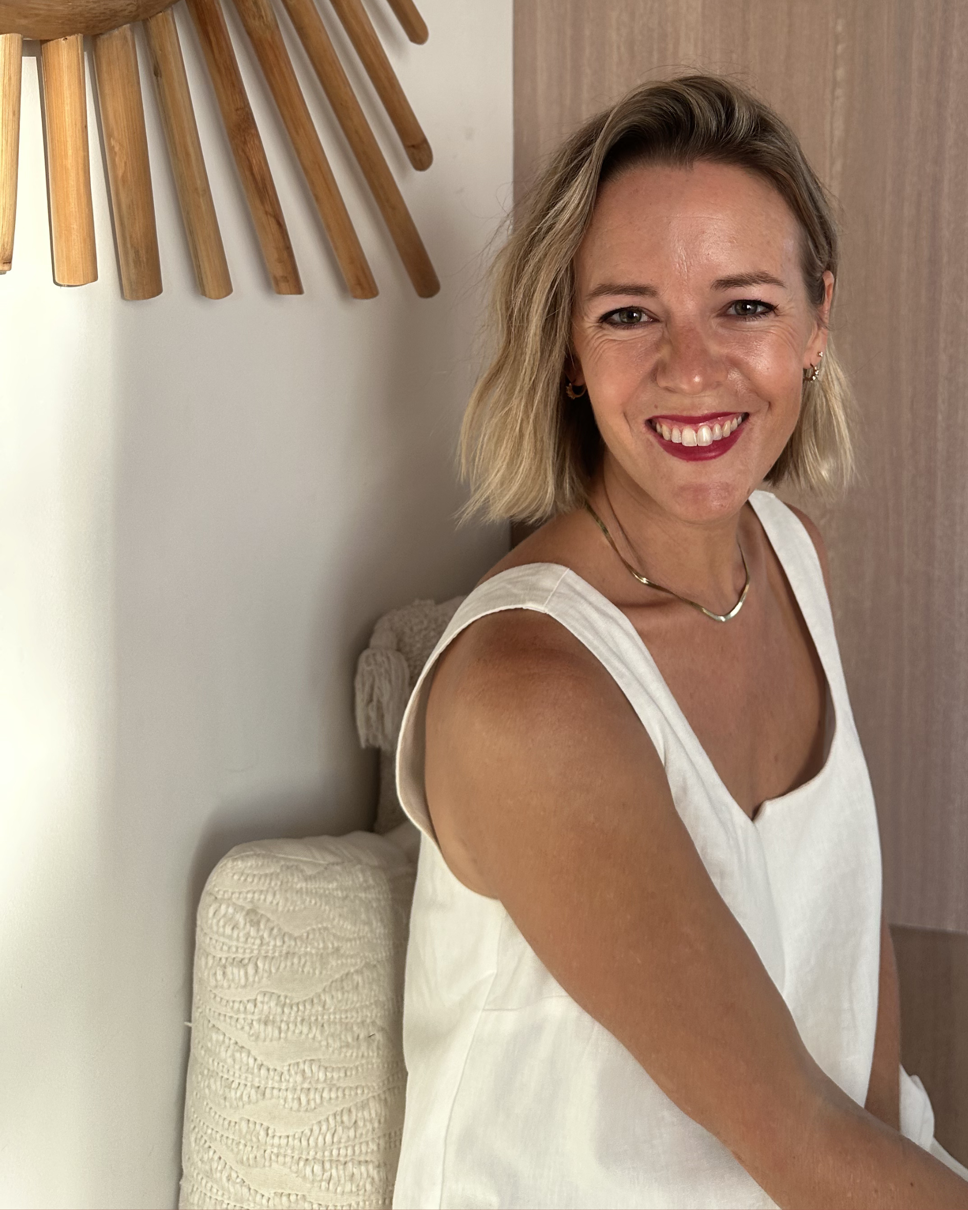 A woman with blonde hair smiling at the camera, sitting on a bed in a bedroom with a wooden headboard and a beige textured pillow in the background.