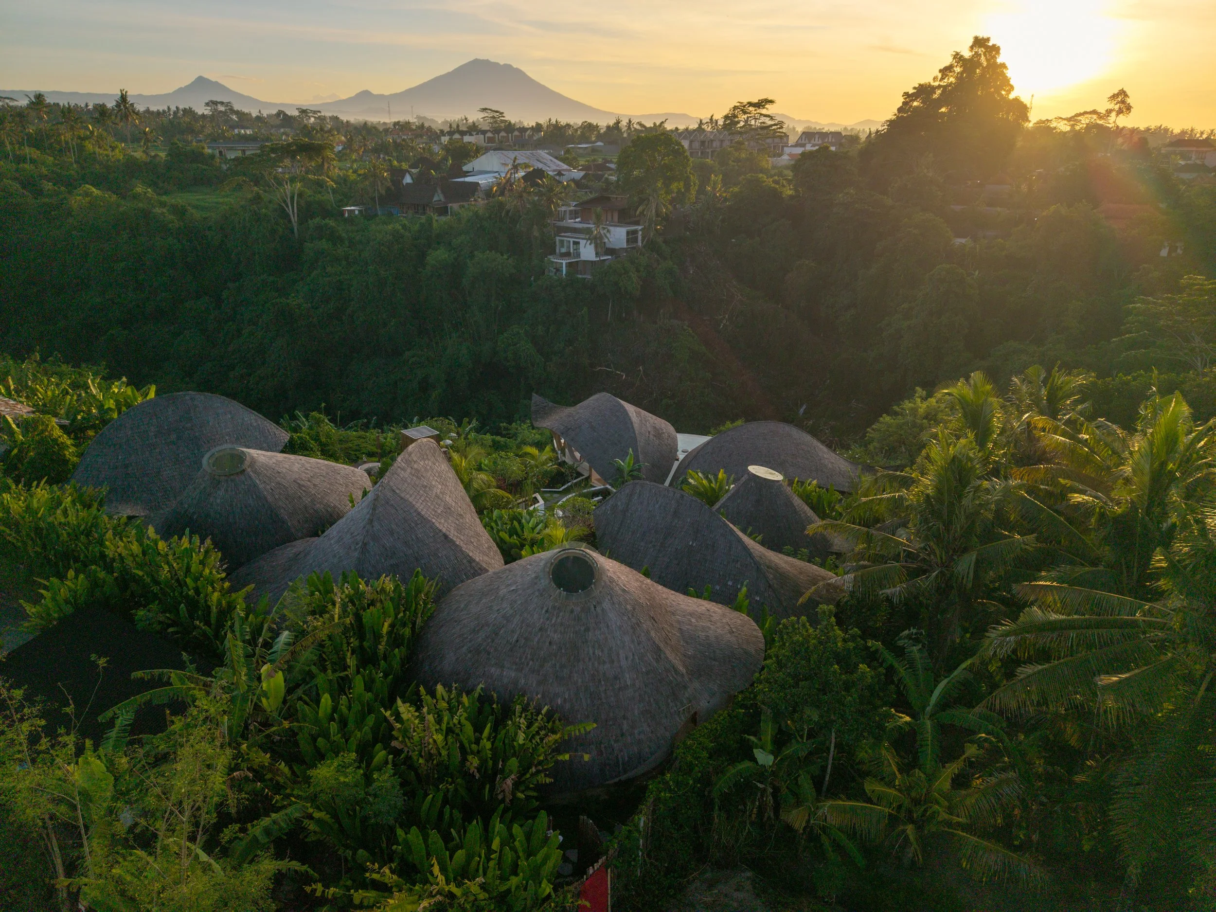 Sunset over a lush green hillside with houses, with mountains in the background and the sun setting on the right.
