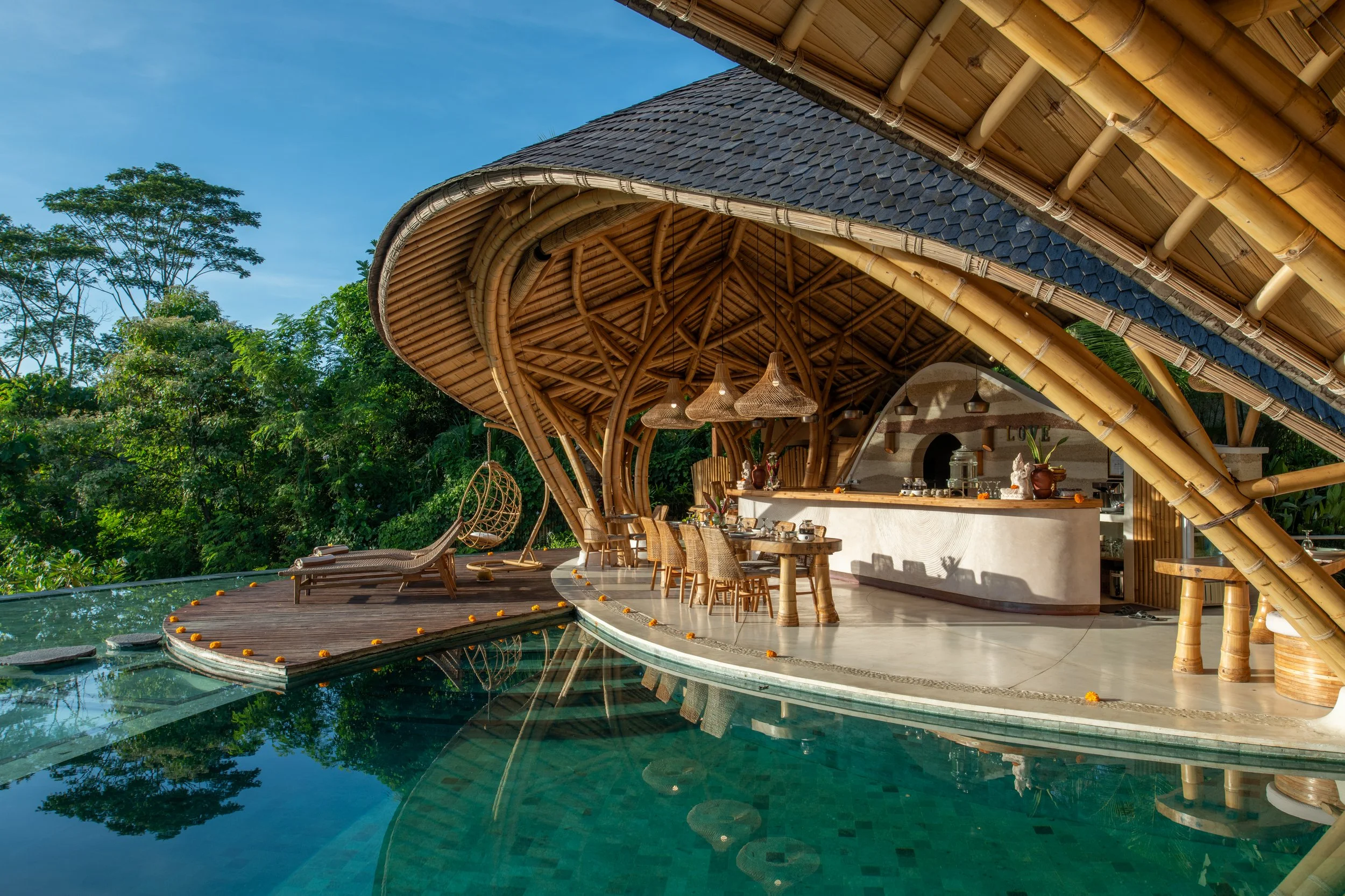 A tropical open-air restaurant or lounge with bamboo and thatch roof, surrounded by lush green trees, with a swimming pool in the foreground, and a blue sky.
