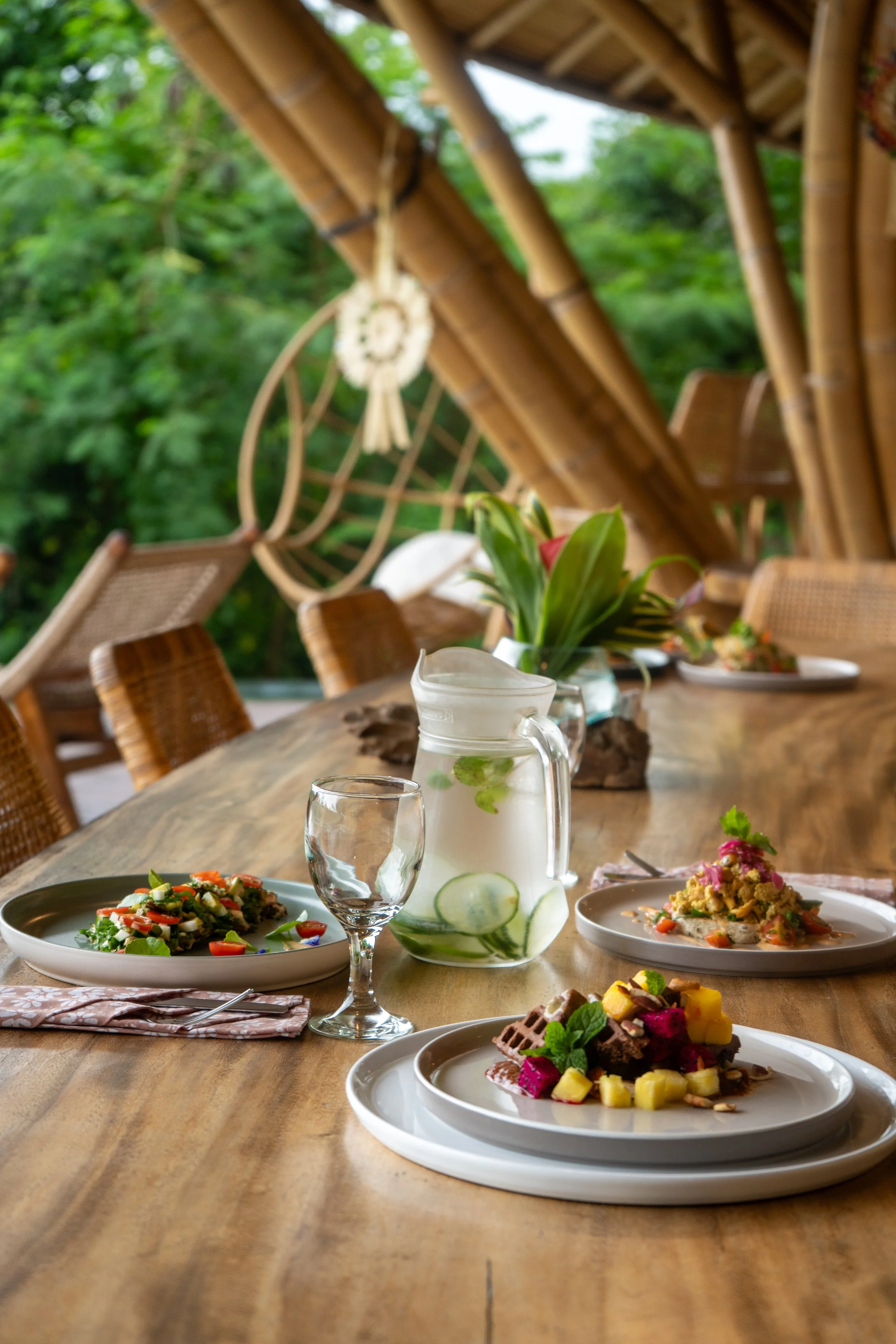 A rustic outdoor dining table set with various dishes, a pitcher of water with cucumber slices, and a vase of lilies, under a bamboo roof with lush greenery outside.