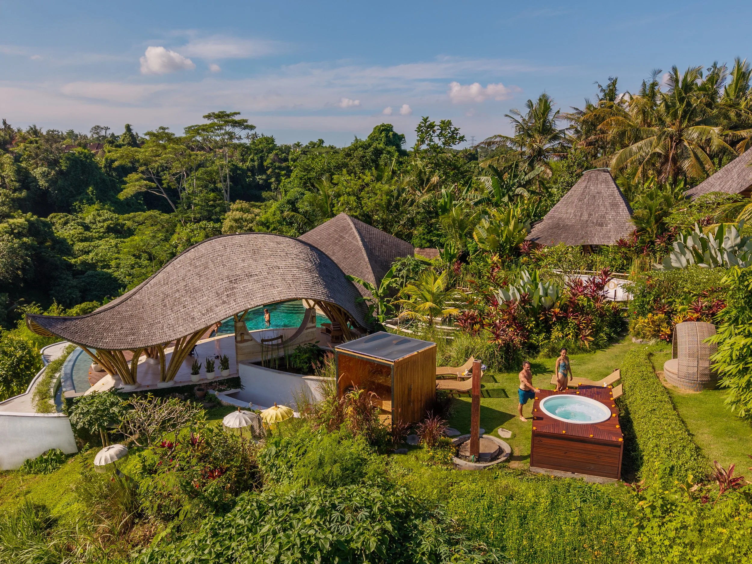 A lush tropical resort with thatched roof structures, a swimming pool, hot tub, and lush green vegetation including palm trees under a blue sky with scattered clouds.