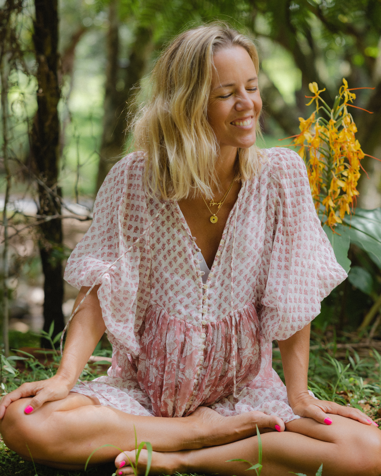 A woman with blonde hair sitting cross-legged outdoors in a forest, smiling with her eyes closed. She is wearing a pink patterned dress, pink nail polish, and layered gold necklaces, including one with the name "Amy." A yellow flowering plant and green trees are in the background.