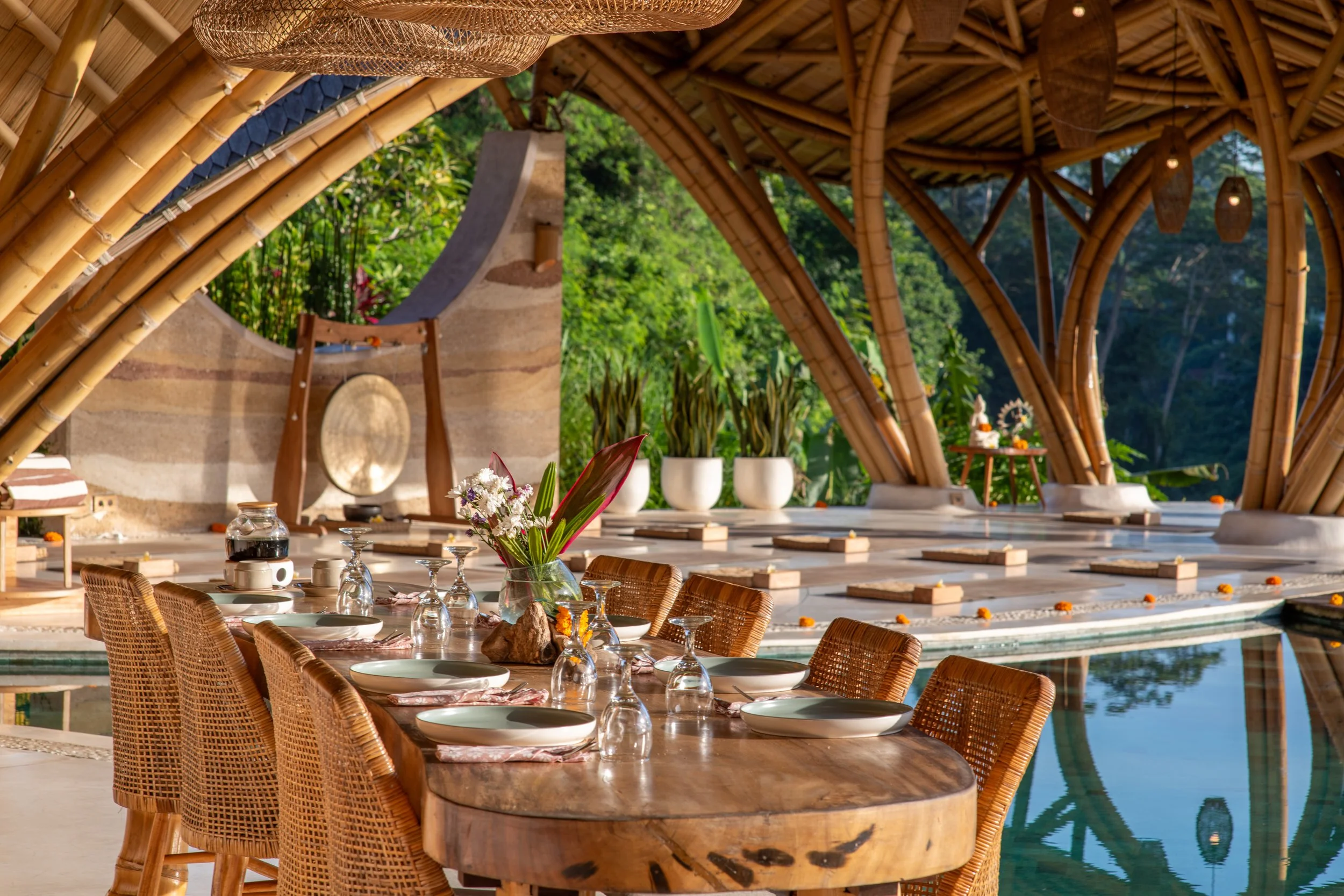 A tropical outdoor dining area with a round table, woven chairs, and decorative items, under a bamboo and woven canopy with lush greenery in the background.