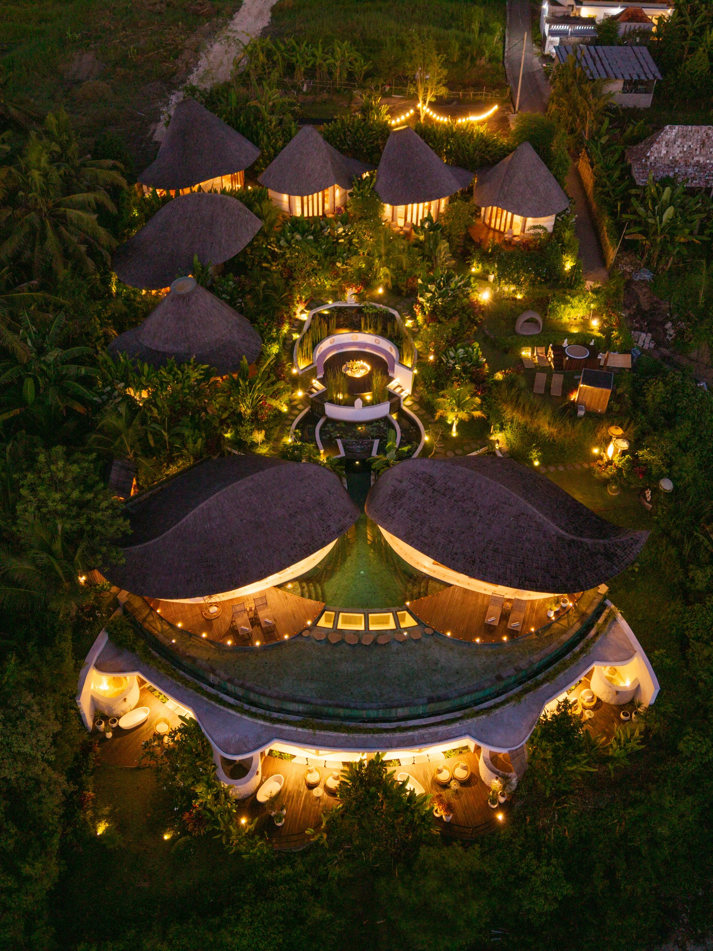 Aerial view of a luxury resort at night featuring circular huts with thatched roofs, lush greenery, and various outdoor seating areas illuminated by warm lighting and string lights.