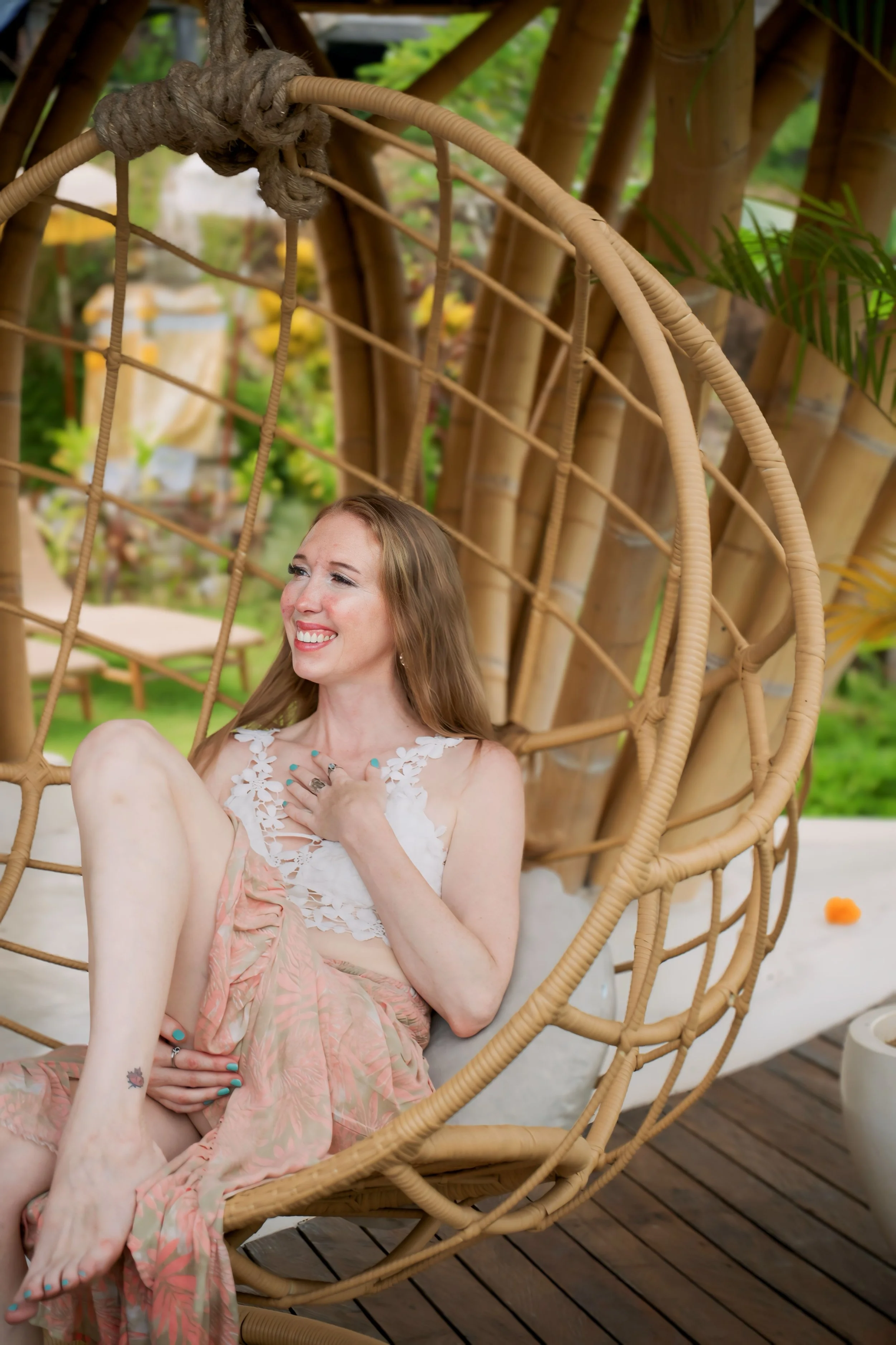 A woman with long hair smiling and sitting in a hanging rattan chair outdoors, surrounded by green plants and bamboo.