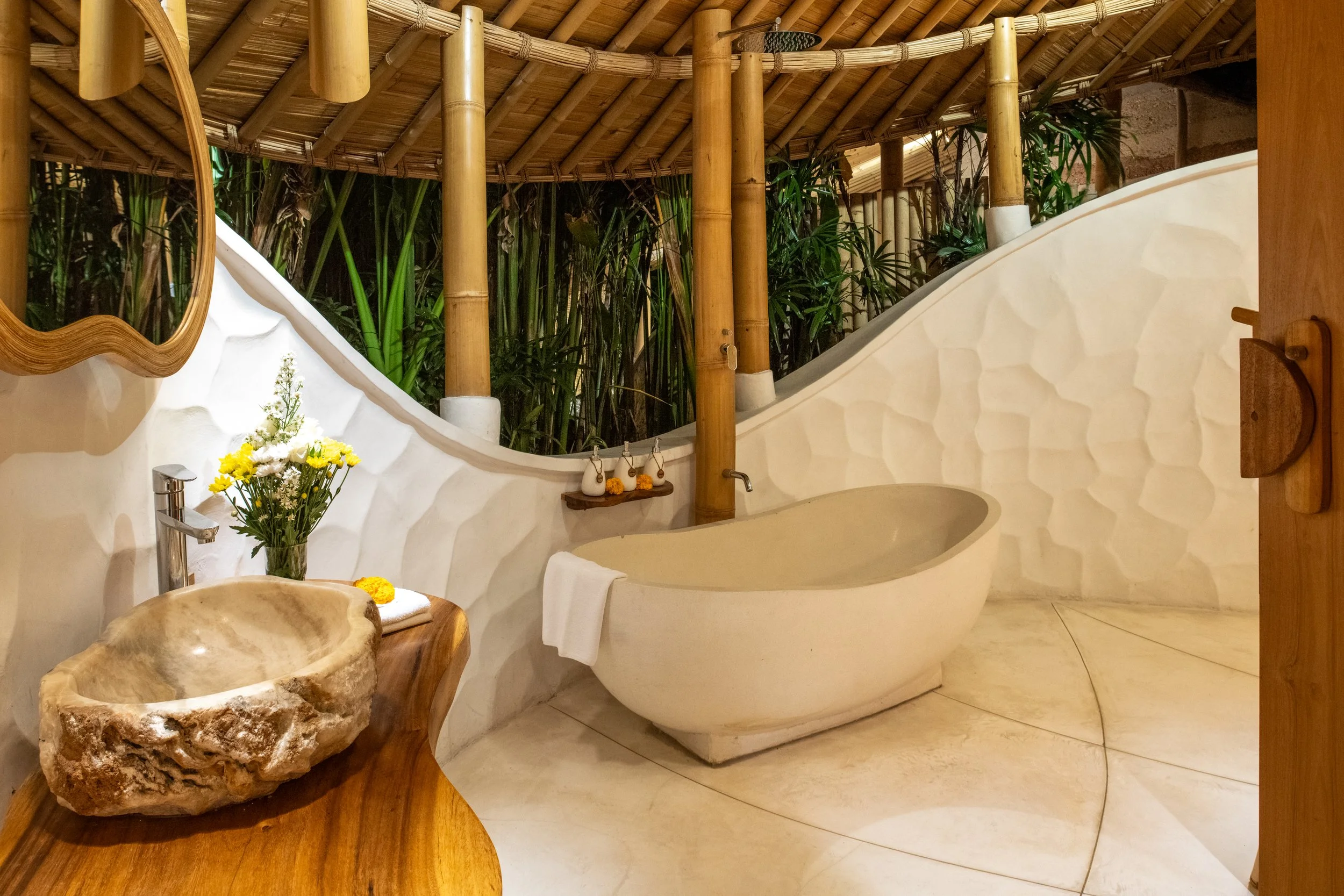 A bathroom with a large, white, oval bathtub next to a curving wall with a textured surface. There is a rustic wooden sink counter made from a natural stone basin and wood, holding a bouquet of white and yellow flowers. The bathroom features bamboo poles and a thatched roof, creating a tropical or spa-like atmosphere. Green foliage is visible behind a bamboo partition.