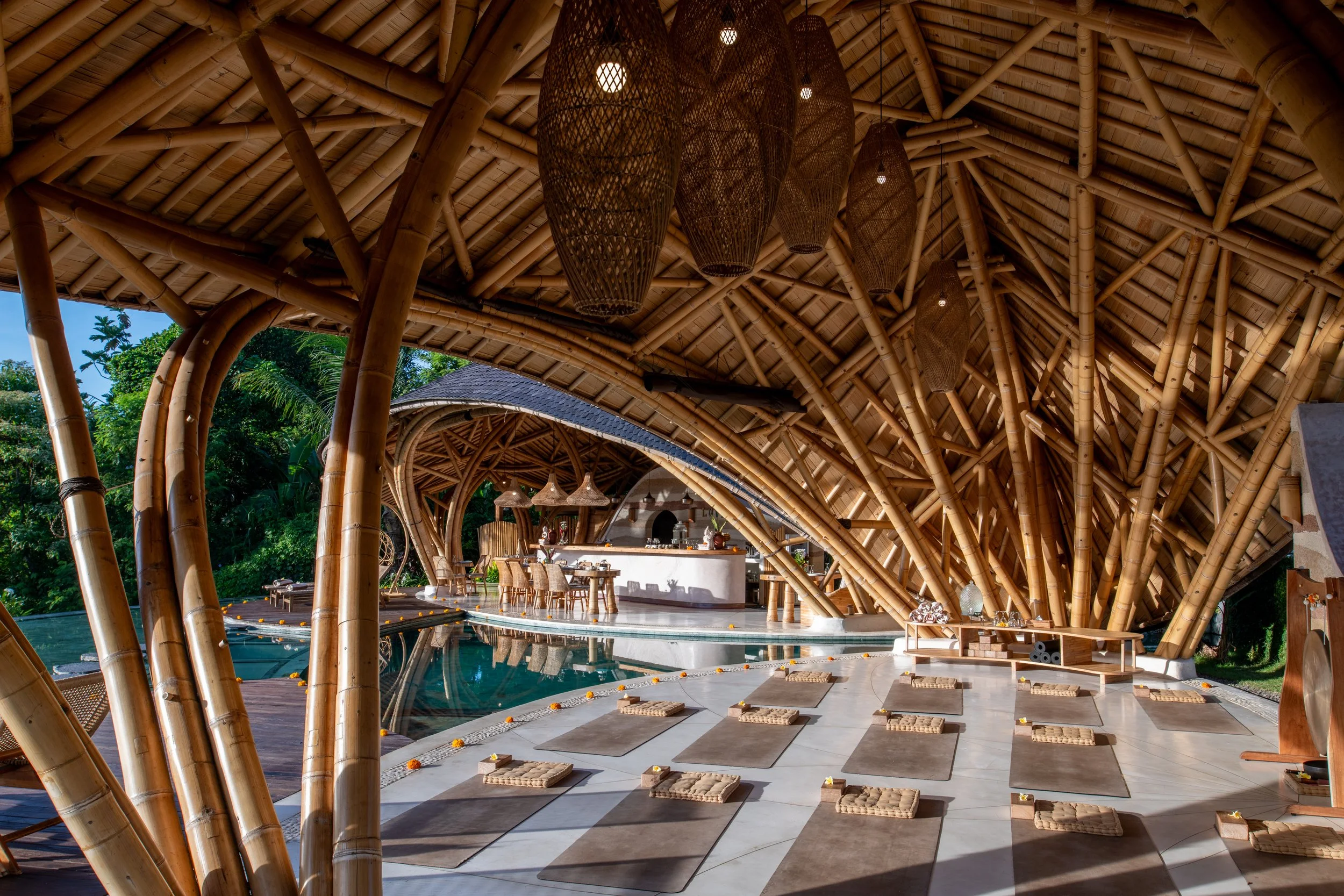 Tropical open-air restaurant with bamboo and thatch architecture, outdoor seating, a swimming pool, and lush greenery in the background.