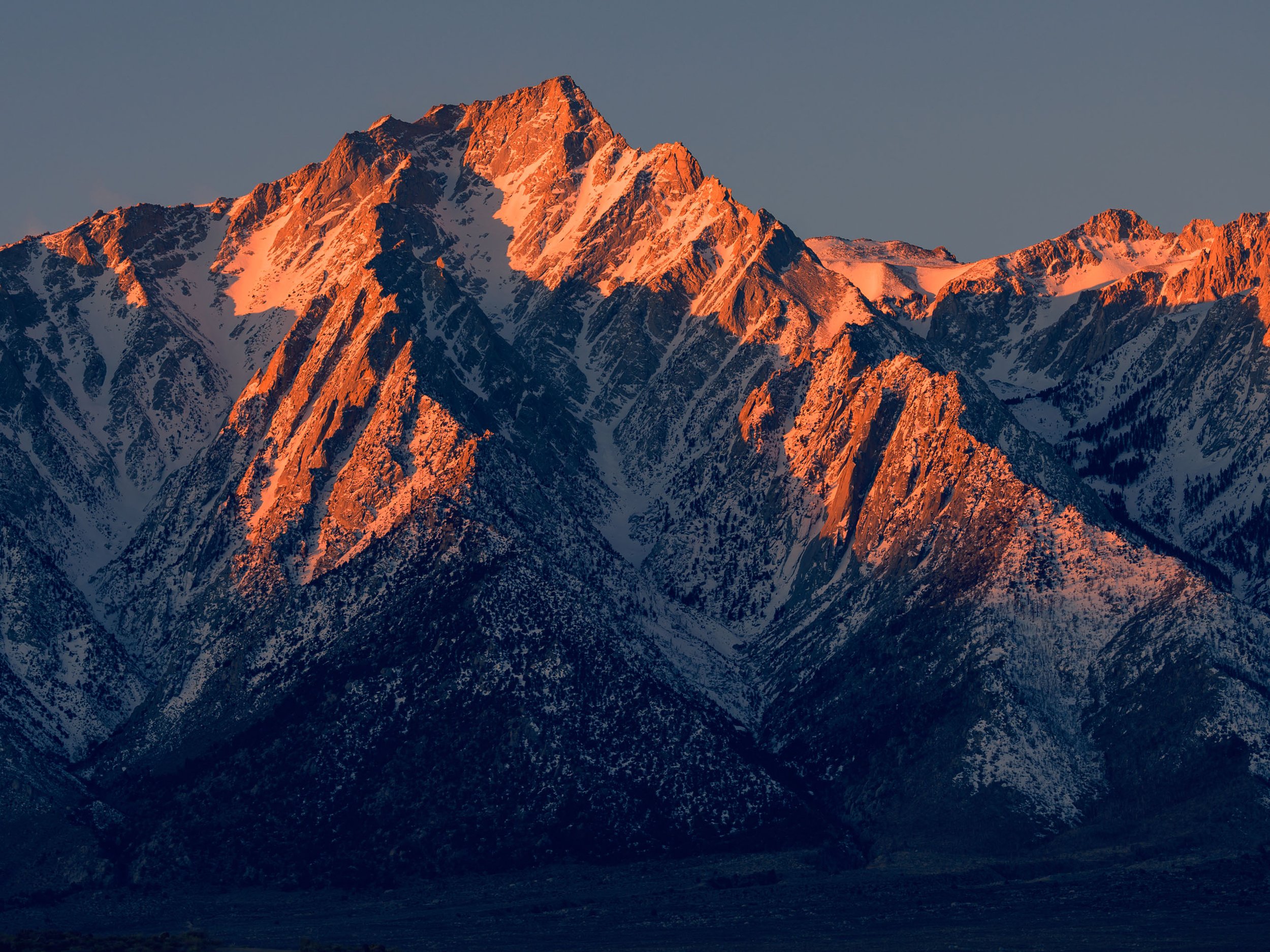 Sunset illuminating snow-capped mountain peaks with a dark valley in the foreground.