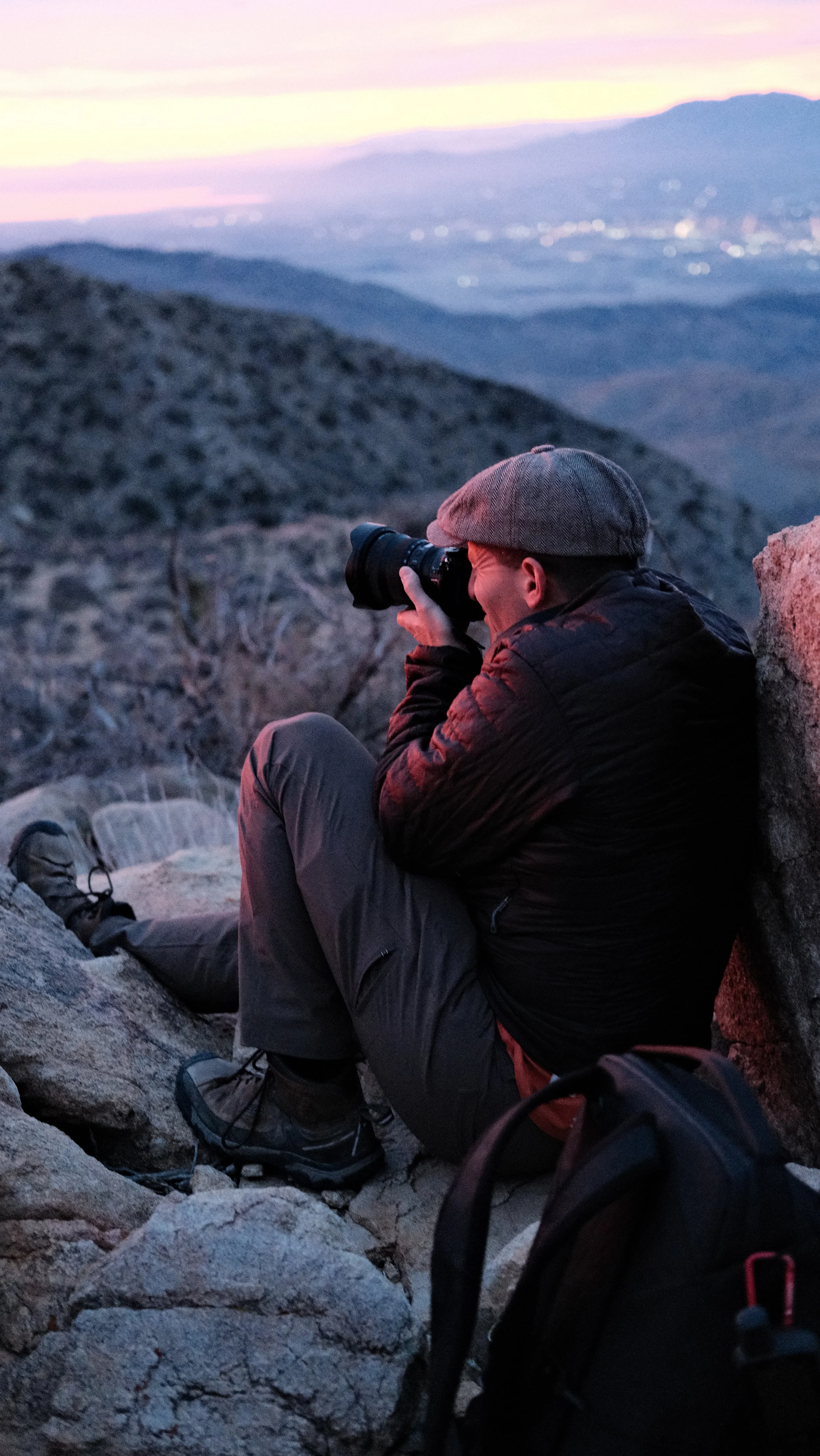 Person sitting on rocky terrain taking photos with a camera during sunrise in a mountainous area.