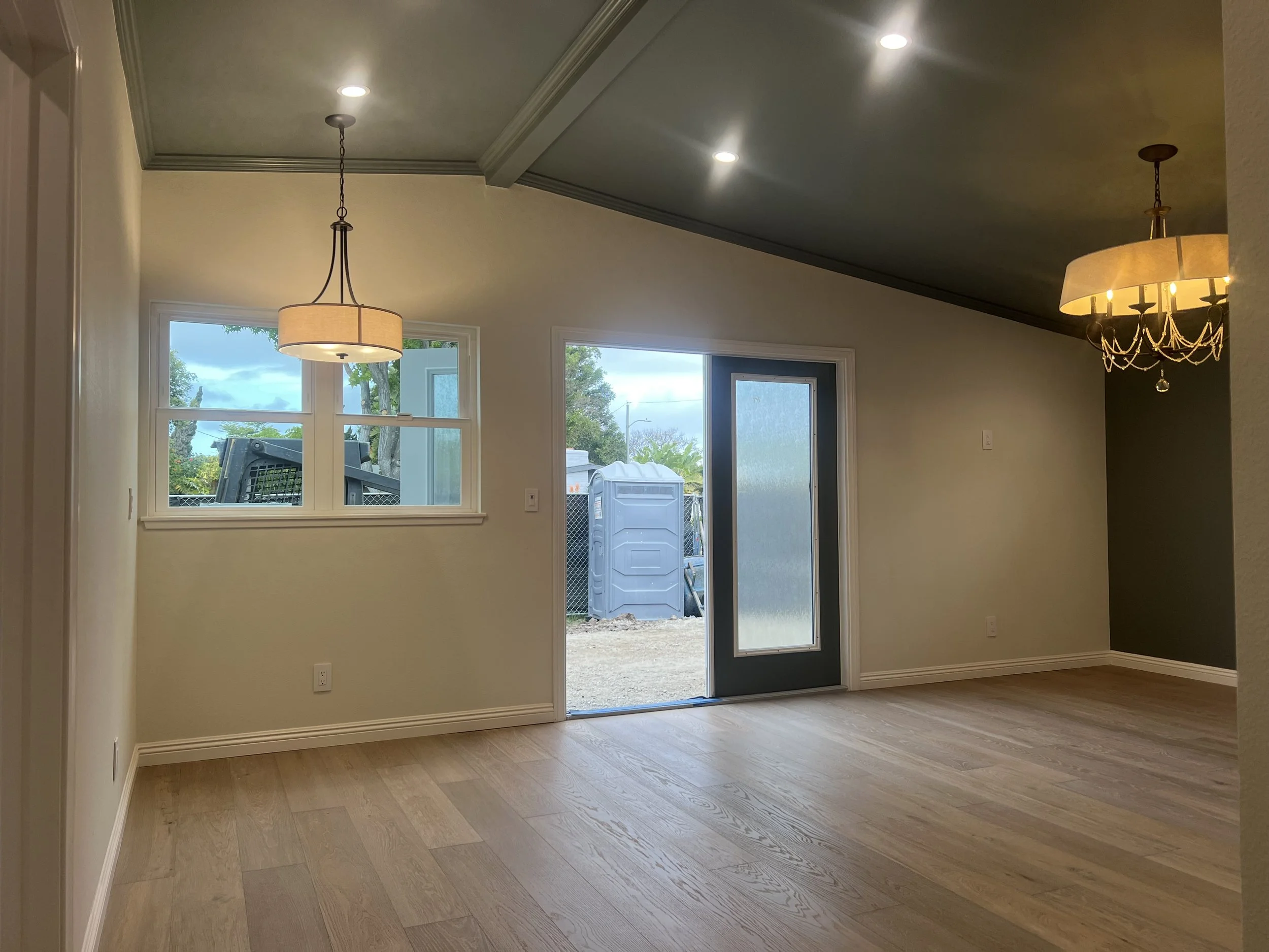 Empty living room with wood flooring, white walls, multiple windows, and two hanging light fixtures, with a door leading outside.