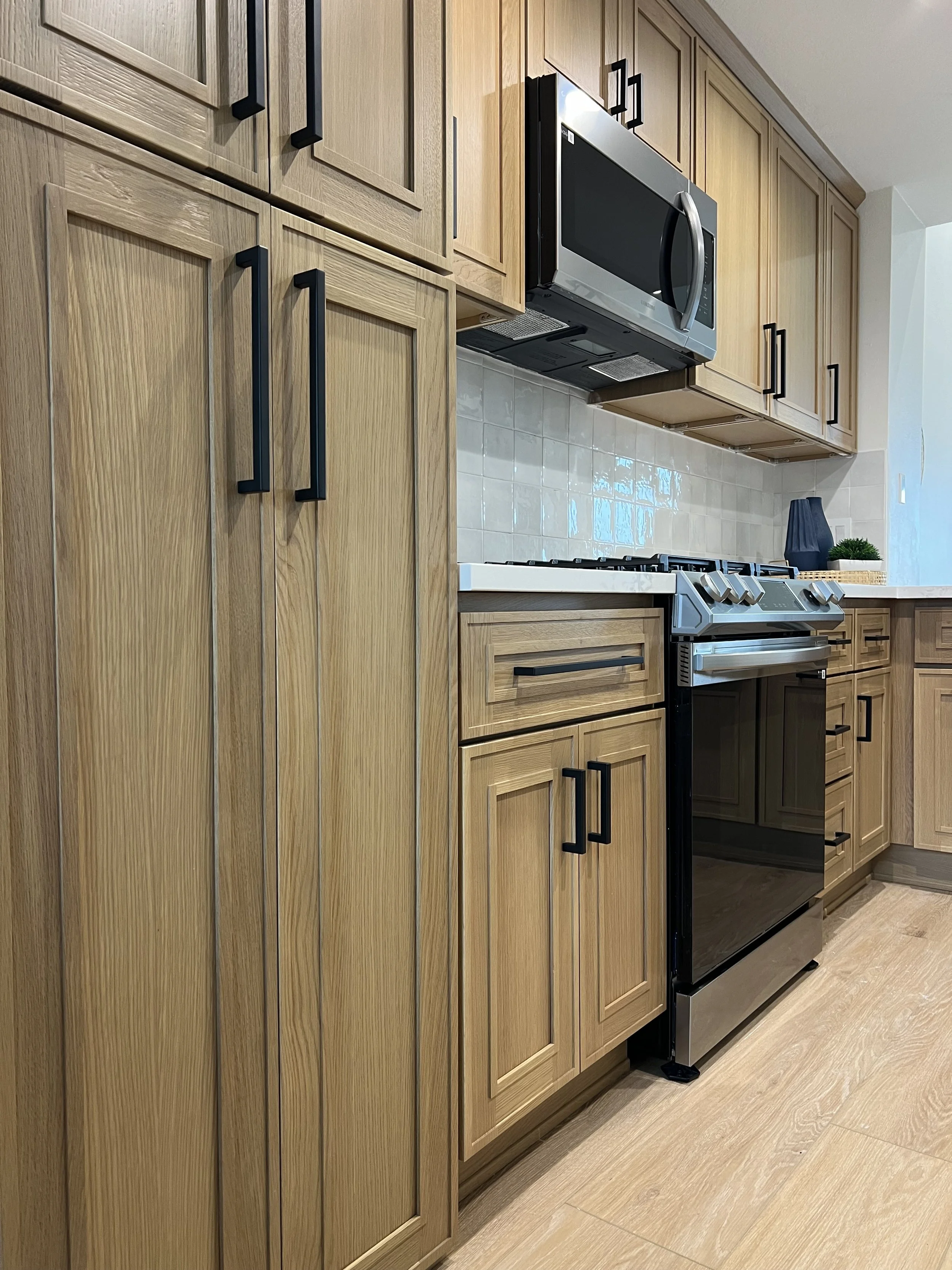 Kitchen with light wood cabinets, black handles, stainless steel microwave, gas stove, beige tile backsplash, and a black vase with a potted plant on the countertop.