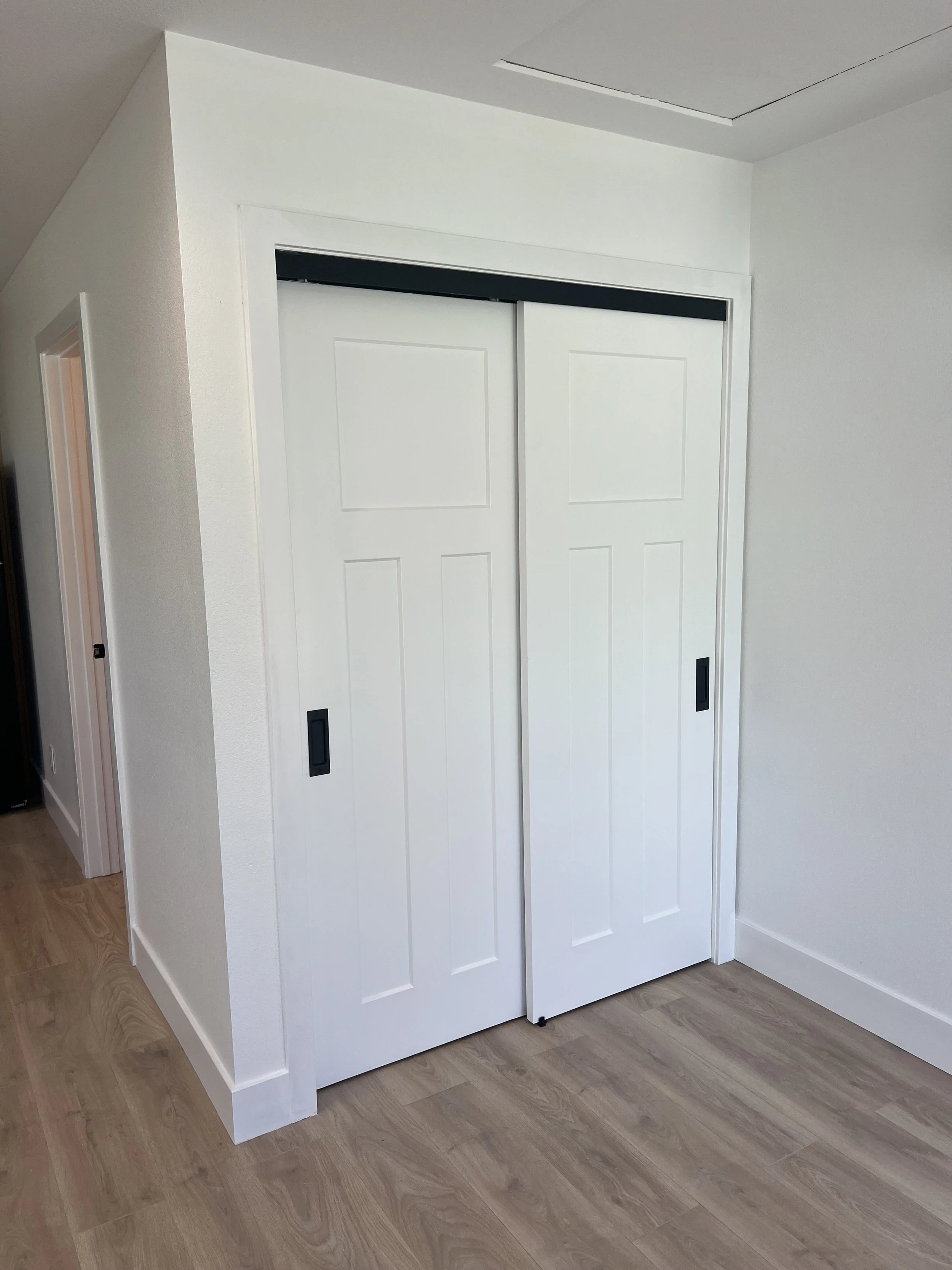 White sliding closet doors with black handles in a corner of a room with wooden floor and white walls.