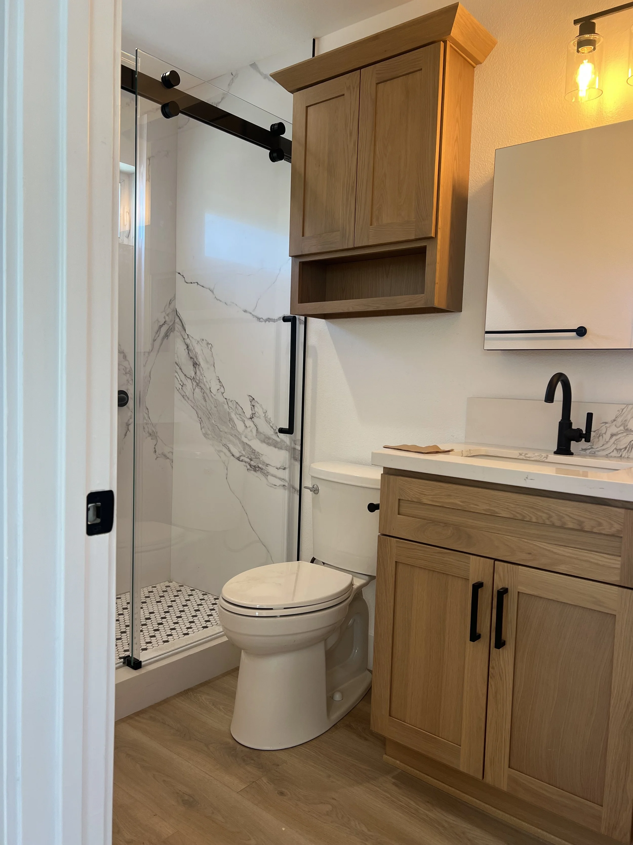 A modern bathroom with a wooden vanity, a marble countertop, and a black faucet. There is a toilet next to the vanity and a shower with a glass door and marble walls. The shower has black fixtures and a mosaic tile floor. A mirror and a wall-mounted 
