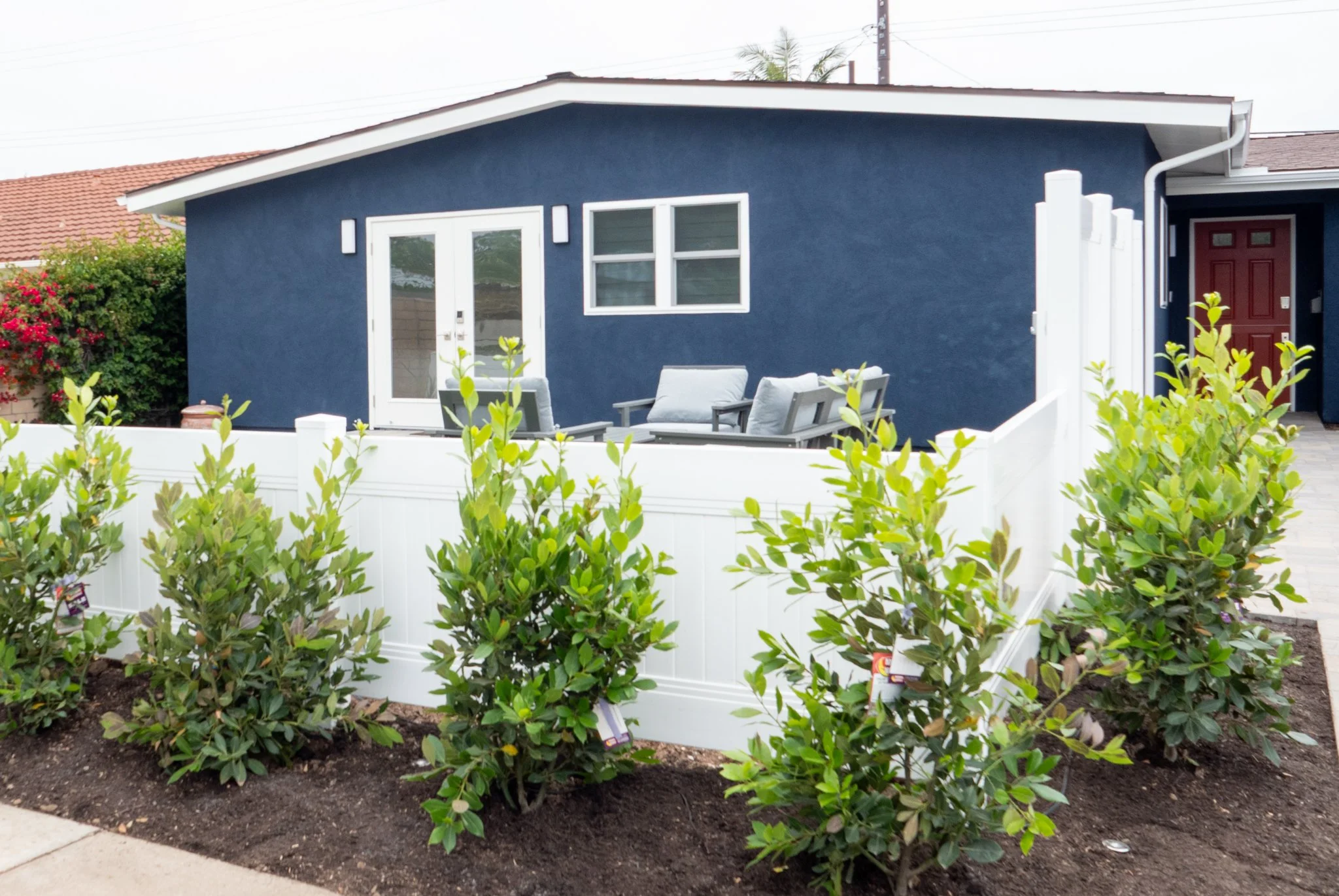 A blue house with white trim and a red front door, outdoor seating area with chairs, surrounded by greenery and plants, with a white fence.