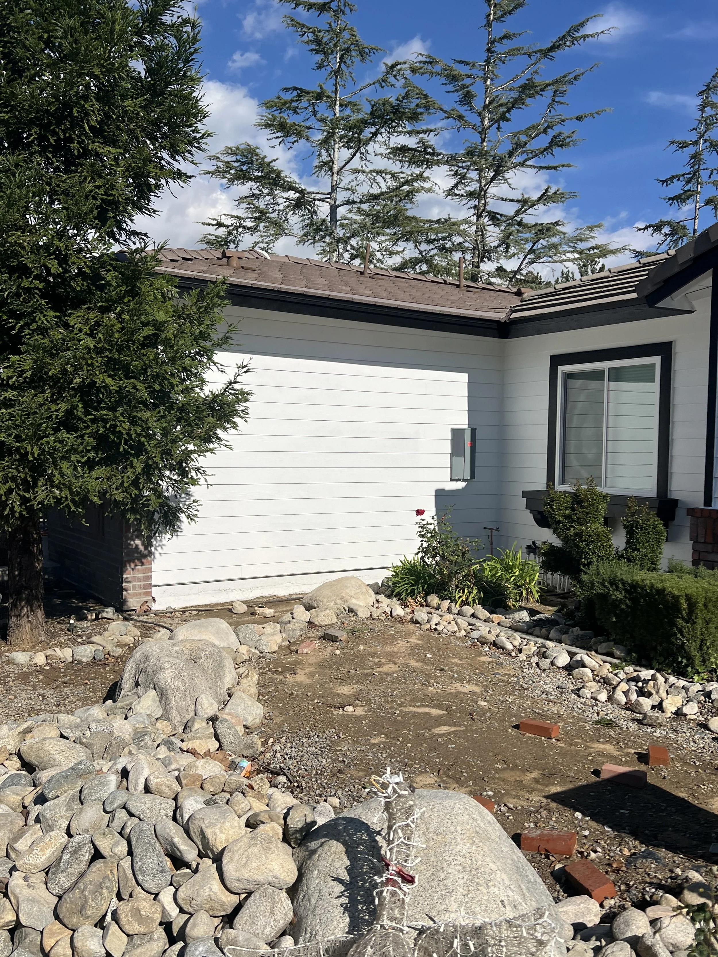 A house with a white exterior, black window frames, and a brown tiled roof. There is a small garden with bushes and flowers, and a rocky yard with a mesh-wire sculpture of an animal, likely a dog, in the foreground. Tall trees and a partly cloudy blu