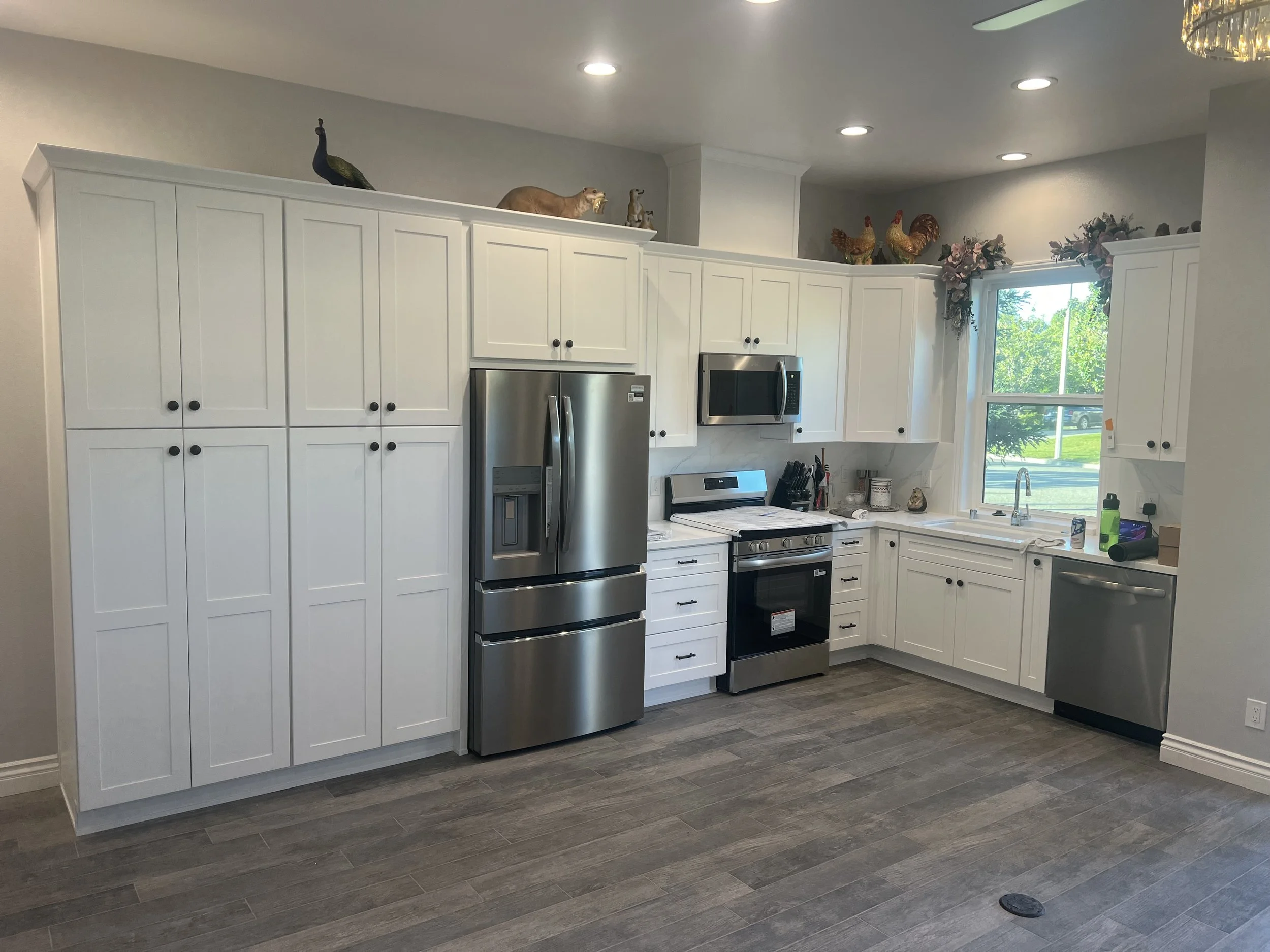A modern kitchen with white cabinets, stainless steel appliances, and a window with greenery outside. Decorative animal figurines are placed on top of the cabinets, including a peacock, lions, and chickens, along with a floral arrangement.