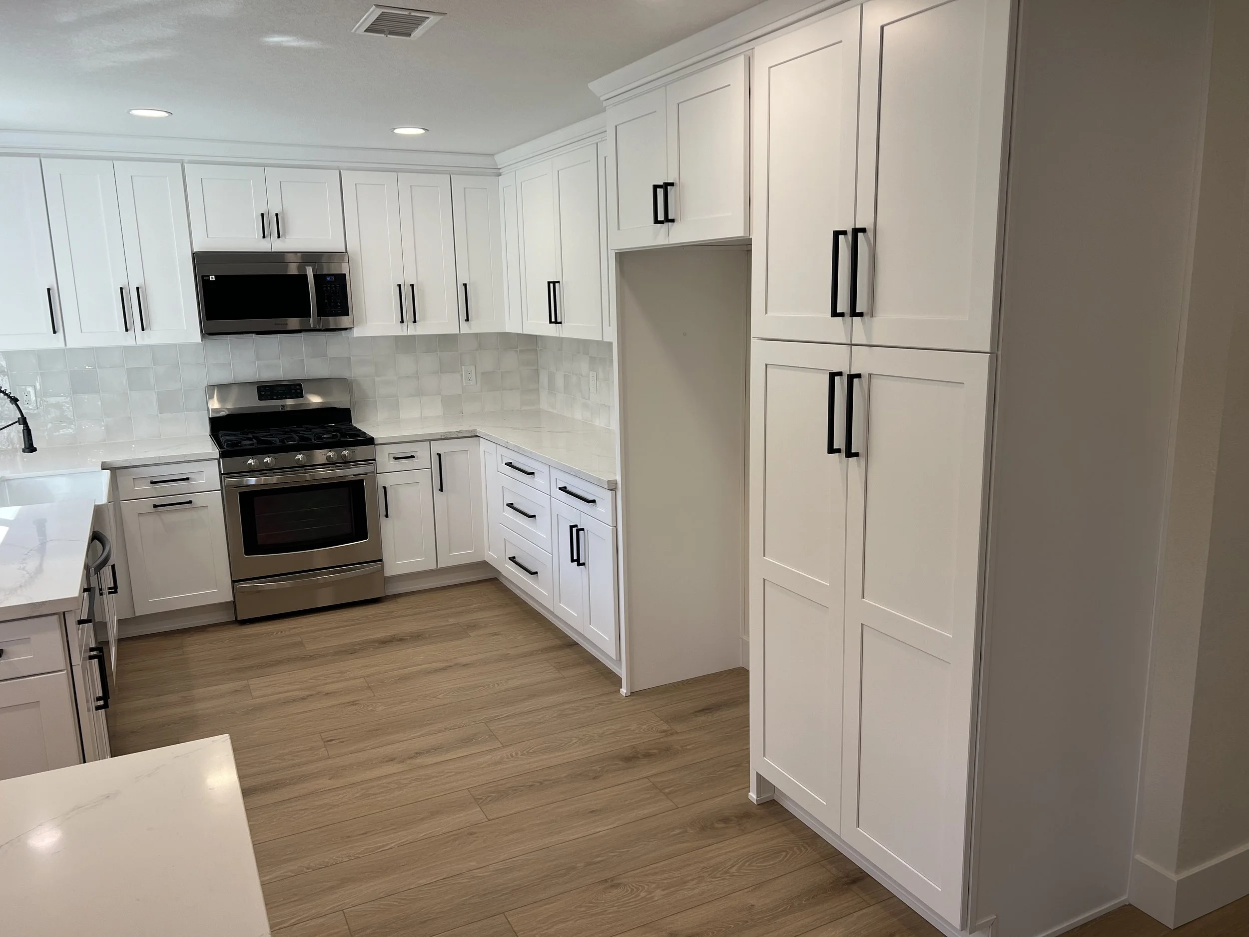 Modern kitchen with white cabinets, stainless steel appliances, black handles, light wood flooring, and a white countertop.