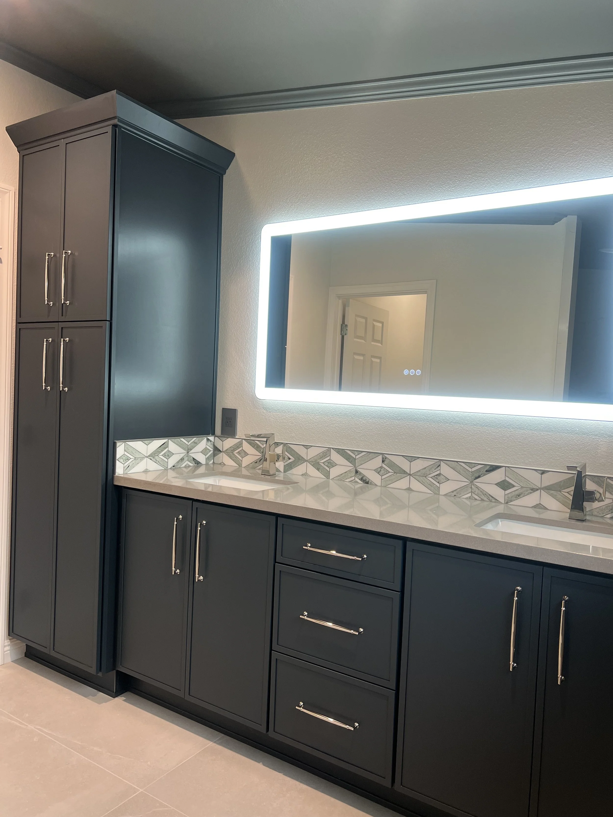 Dark blue bathroom vanity with multiple cabinets and drawers, a sink, a large illuminated mirror, and a patterned backsplash, with beige floor tiles.