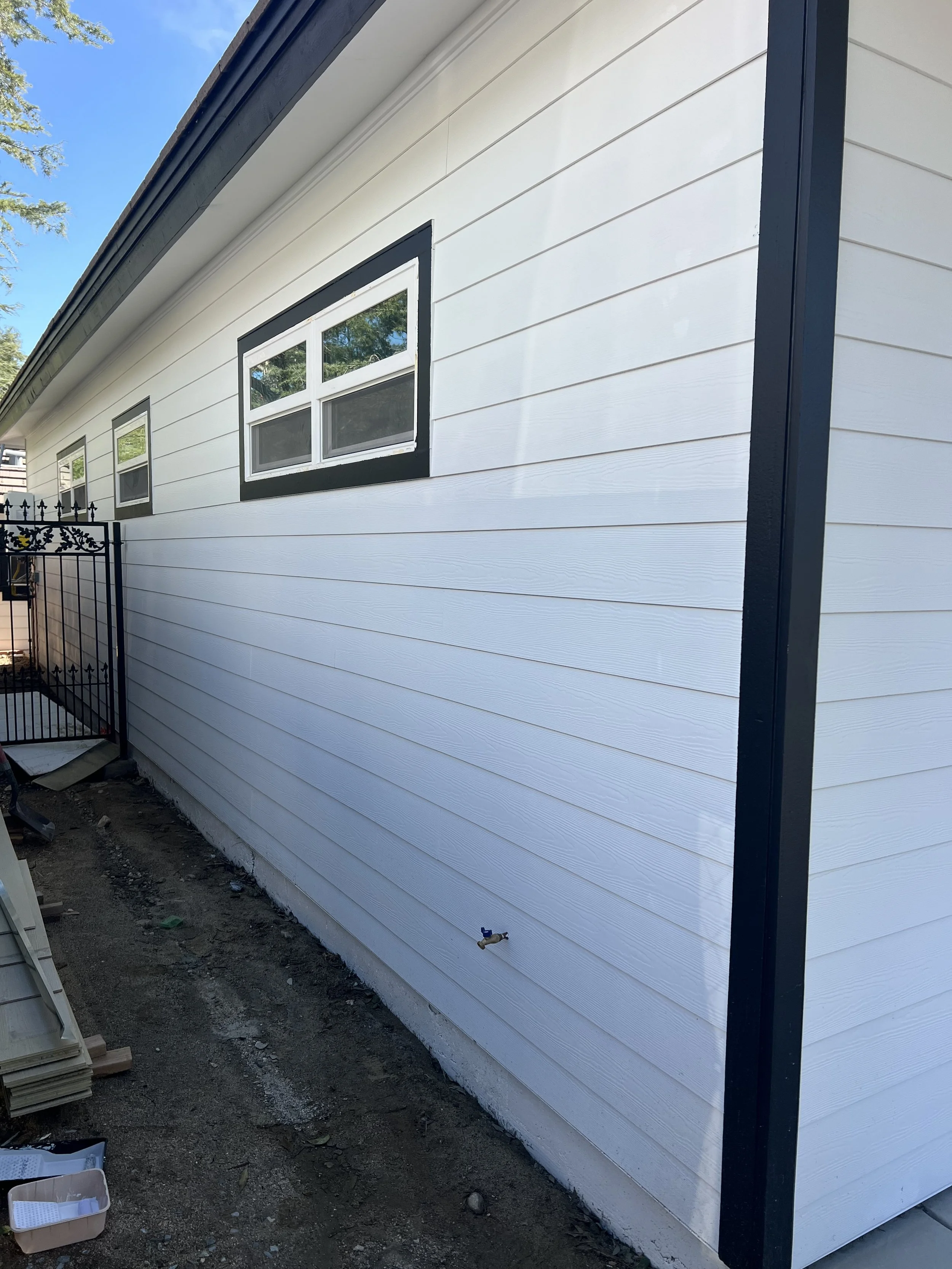 A house siding with white horizontal panels, black window frames, and black trim on the corner. Construction materials and dirt are visible on the ground below.