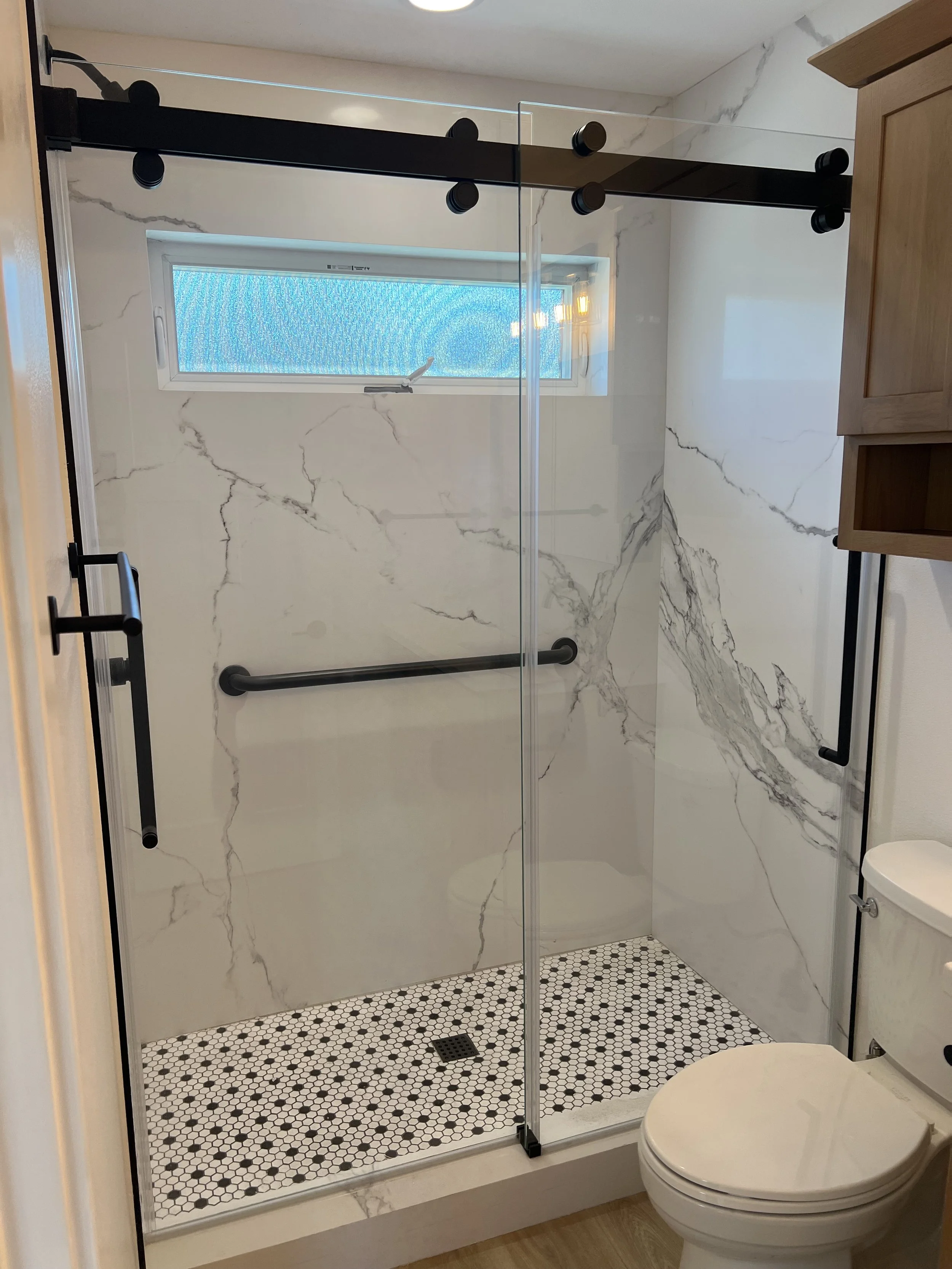 Modern bathroom shower with glass enclosure, white marble walls with gray veining, black sliding barn door hardware, black and white hexagonal tiled floor, small window, and a white toilet in the foreground.
