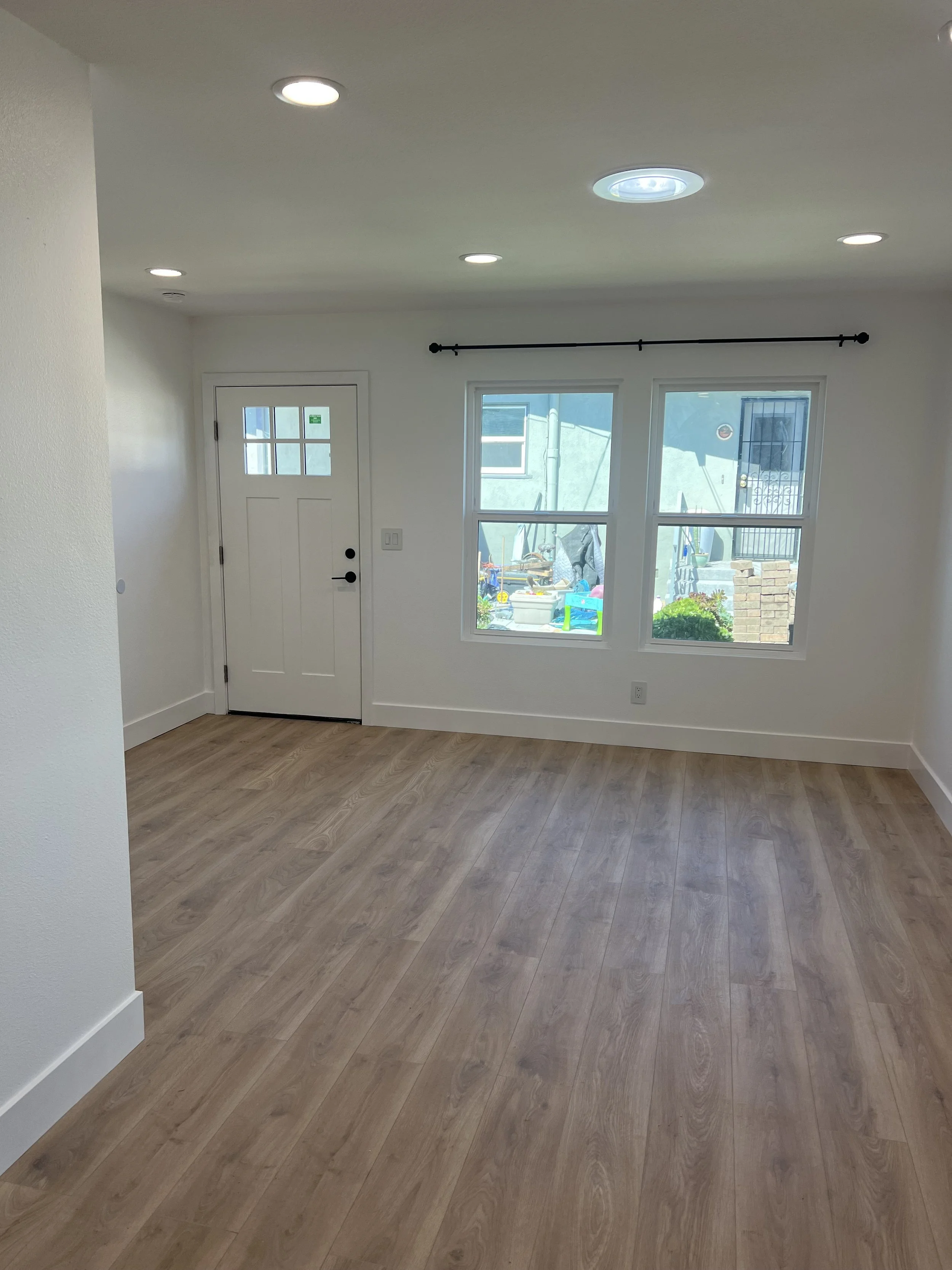 Empty room with wood-look flooring, white walls, a white door with small window panes, and two large windows showing an outdoor scene with a house, plants, and a small brick structure.