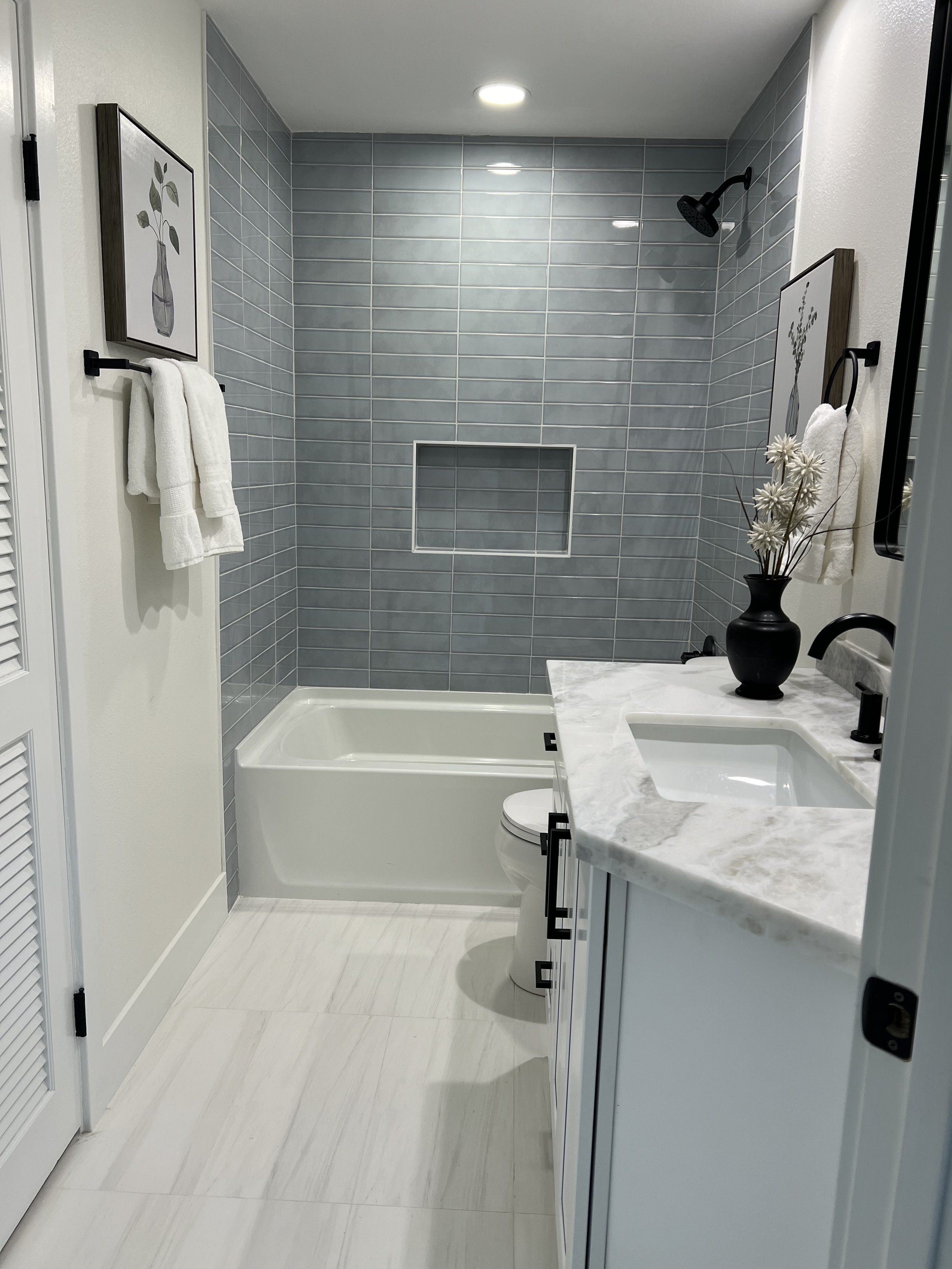 Modern bathroom with light gray tiled shower area containing a built-in niche, white vanity with marble countertop, black fixtures, a black vase with white decorative flowers, and framed artwork on the wall.