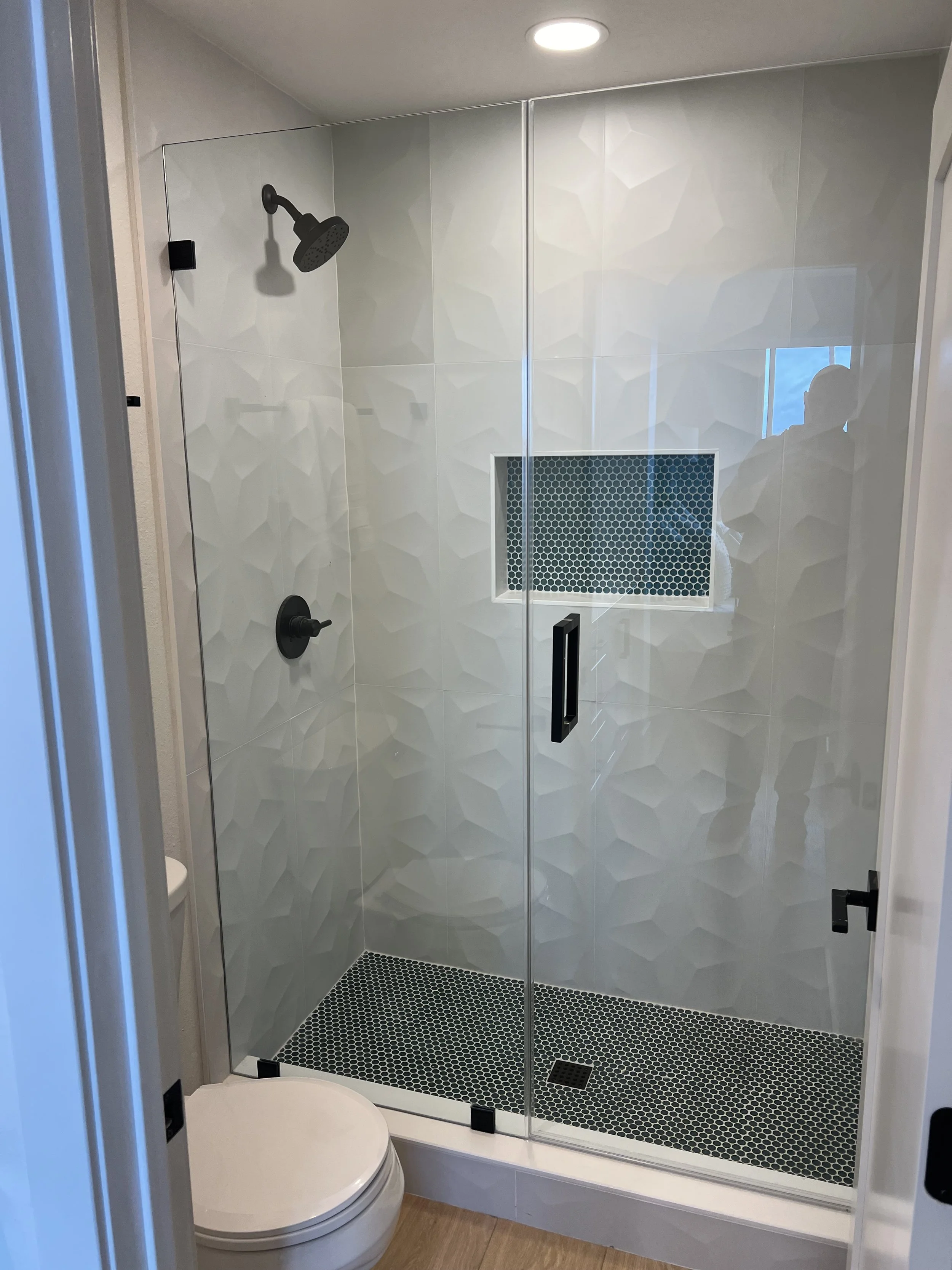 A modern glass-enclosed shower with black fixtures, textured white wall tiles, a built-in soap niche with blue patterned tiles, and a dark hexagon tile floor, adjacent to a white toilet and wood flooring.