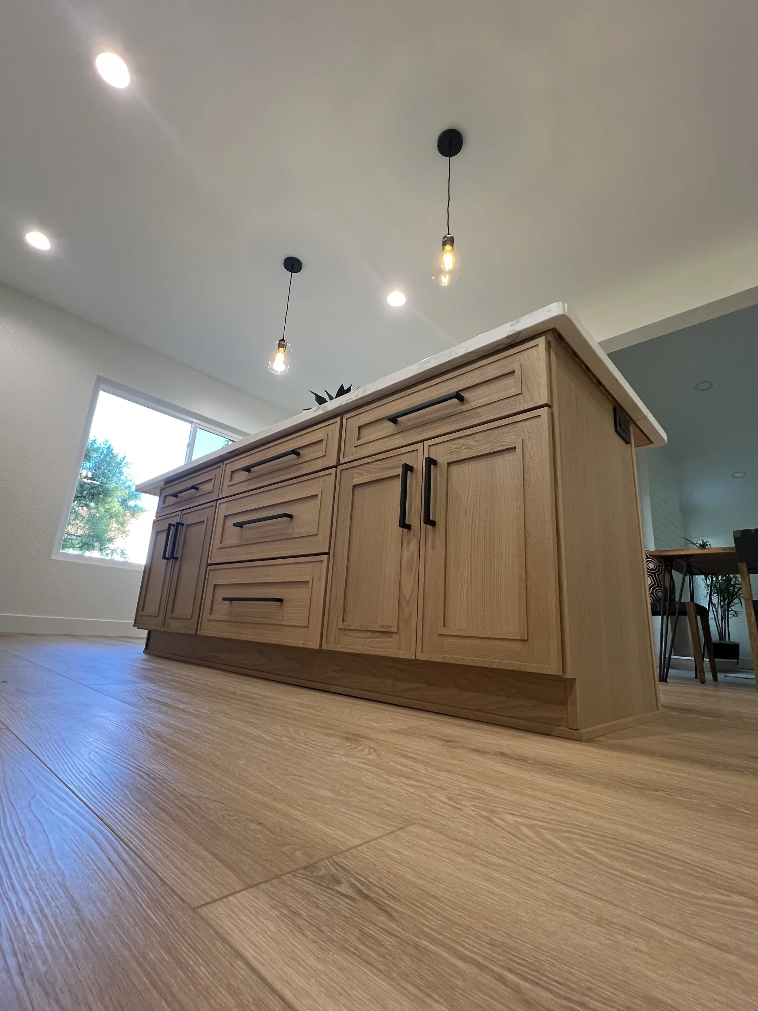 View of a wooden kitchen island with black handles, set in a room with wooden flooring, a window letting in natural light, and ceiling lights.