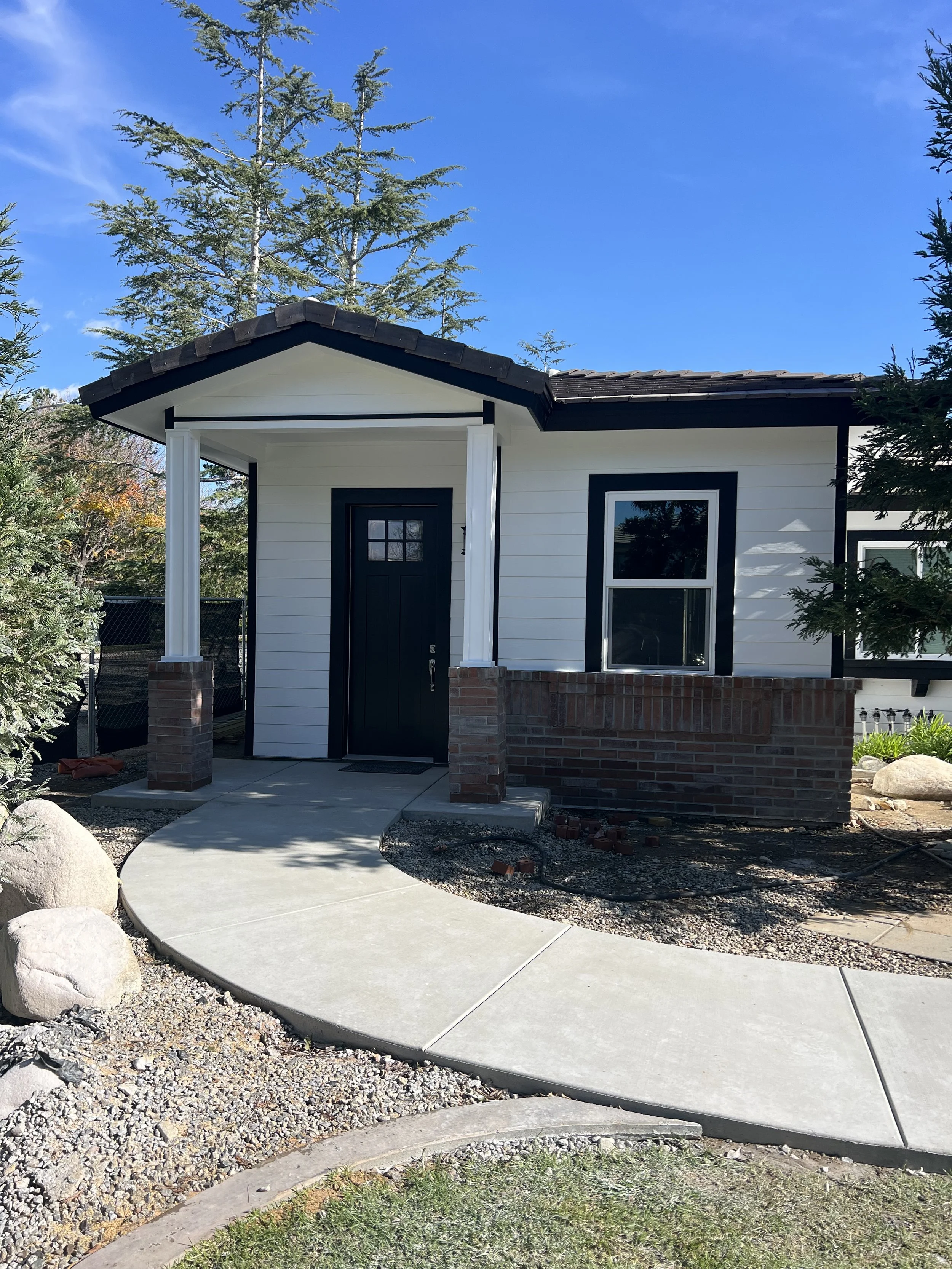 A small white house with black trim and brick accents, a black door, and a concrete pathway in the front yard, surrounded by trees and rocks under a blue sky.