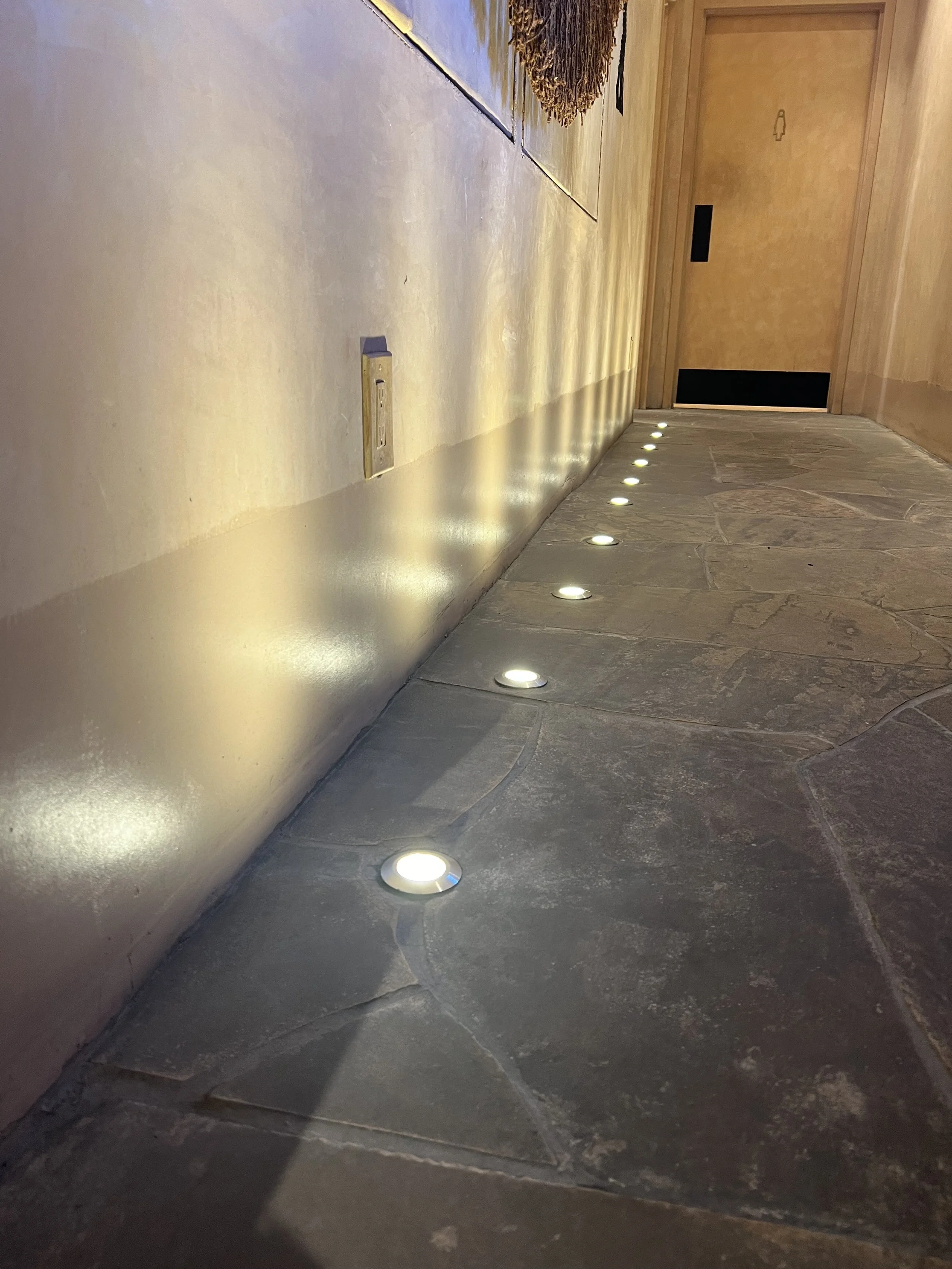 Indoor hallway with a row of small round lights embedded in the floor, a wooden door with a restroom sign, and a stone-tiled section on the wall.