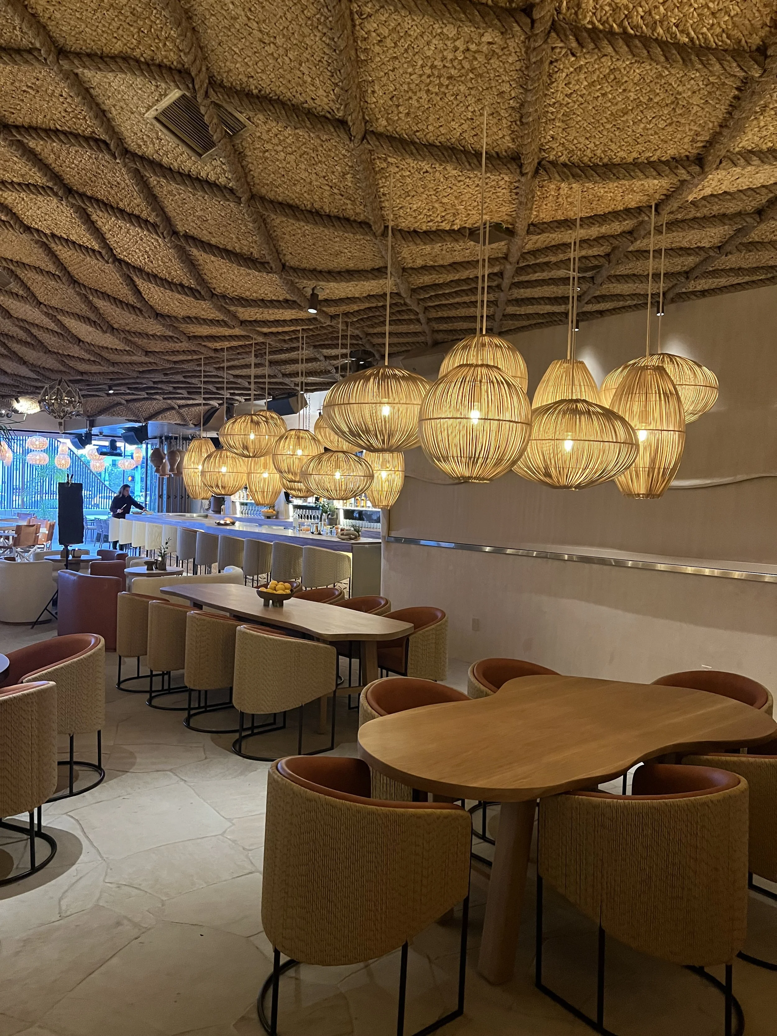 Interior of a restaurant with a woven straw ceiling, hanging paper lanterns, and a bar area in the background. There are round tables with chairs and a person working behind the bar.